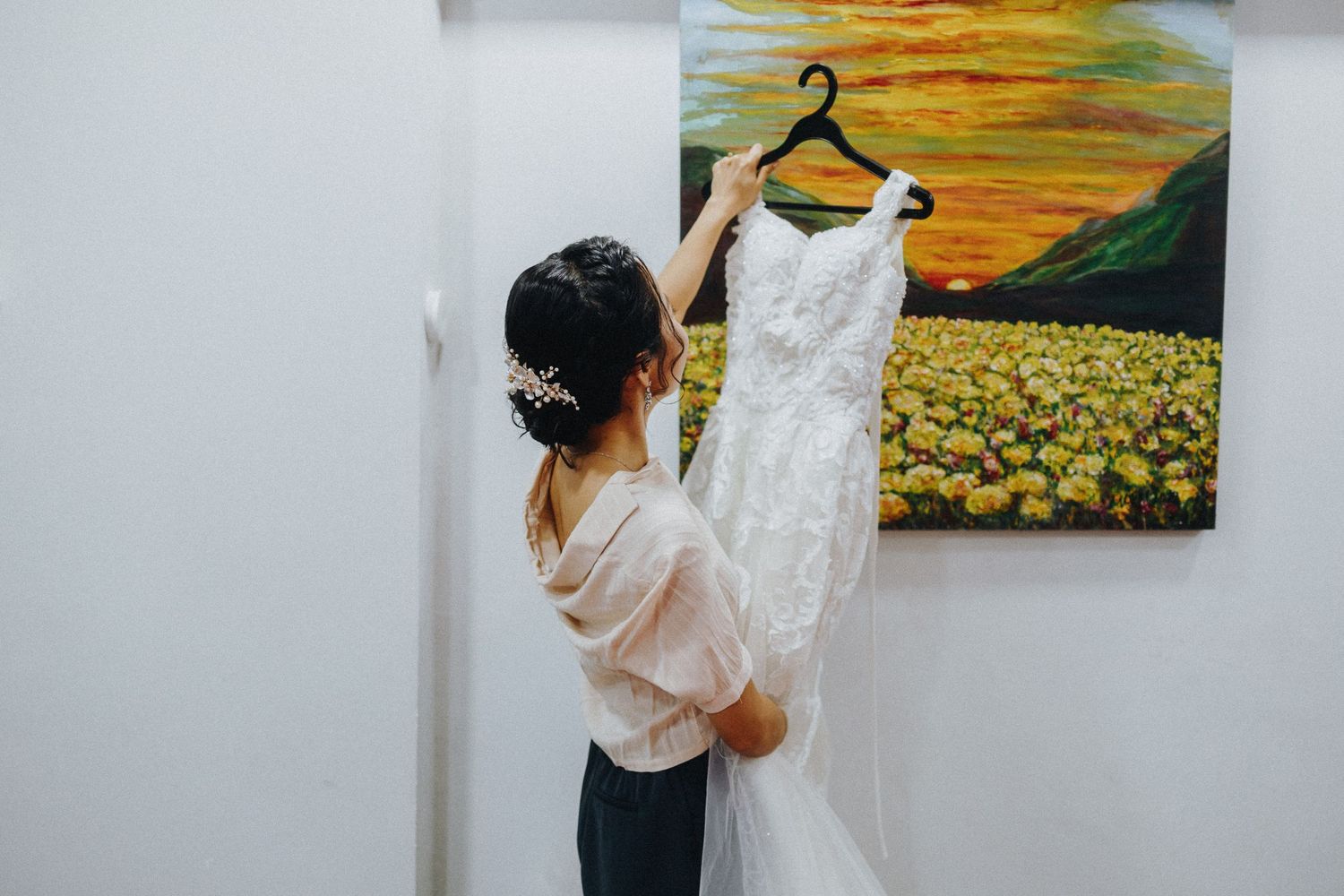 Someone holds up an elegant white lace wedding dress on a hanger against a wall with a vibrant sunflower painting.