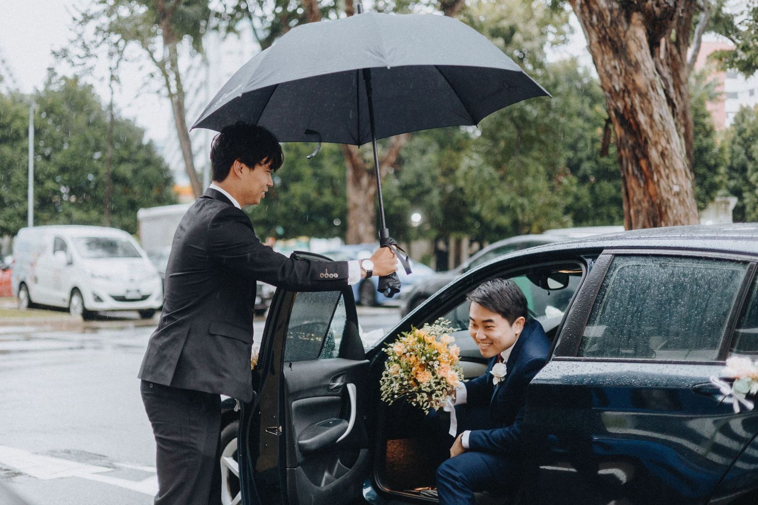 A figure holds a black umbrella while assisting someone into a car on a rainy day.