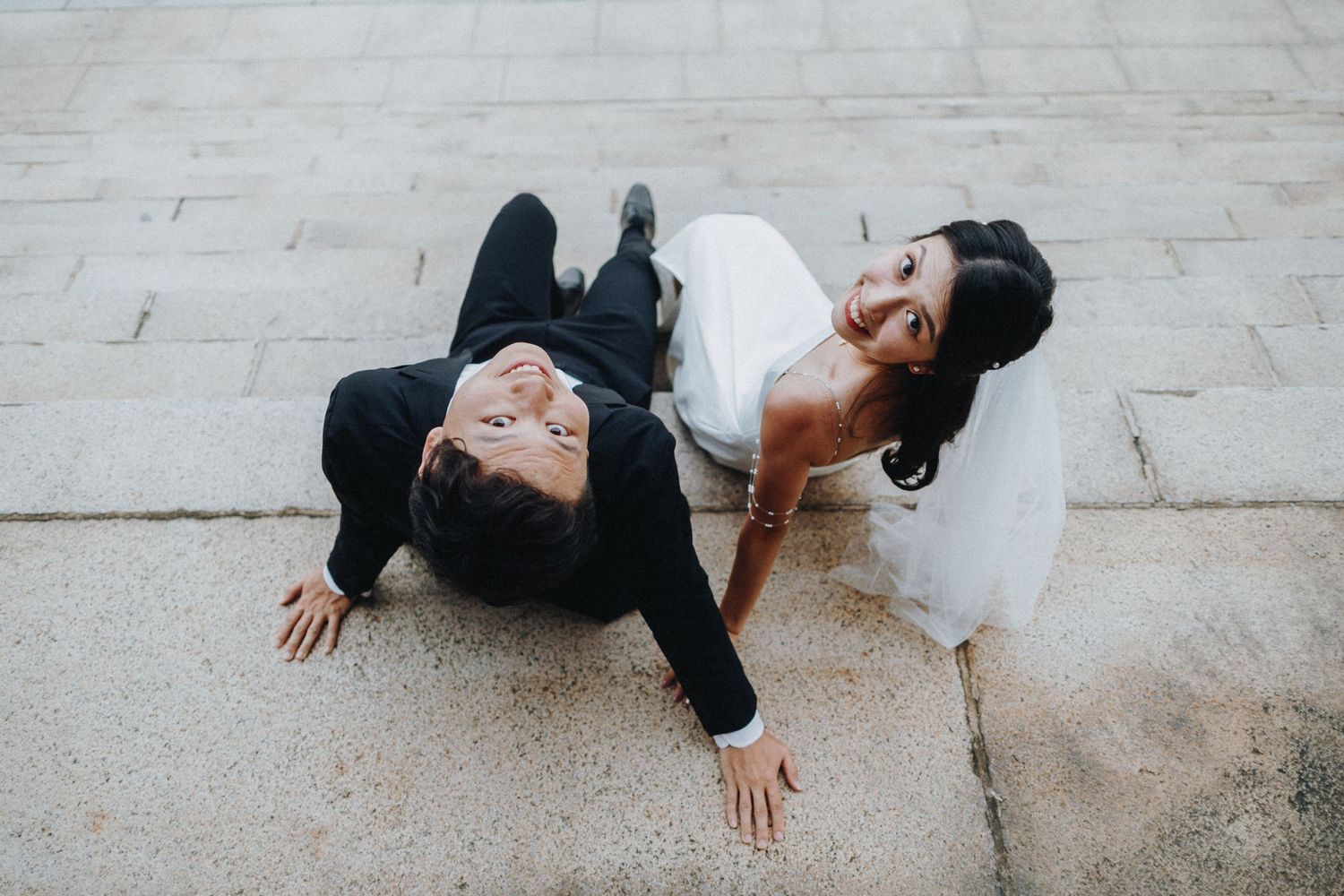 Candid wedding photo with couple lying on ground in formal attire.