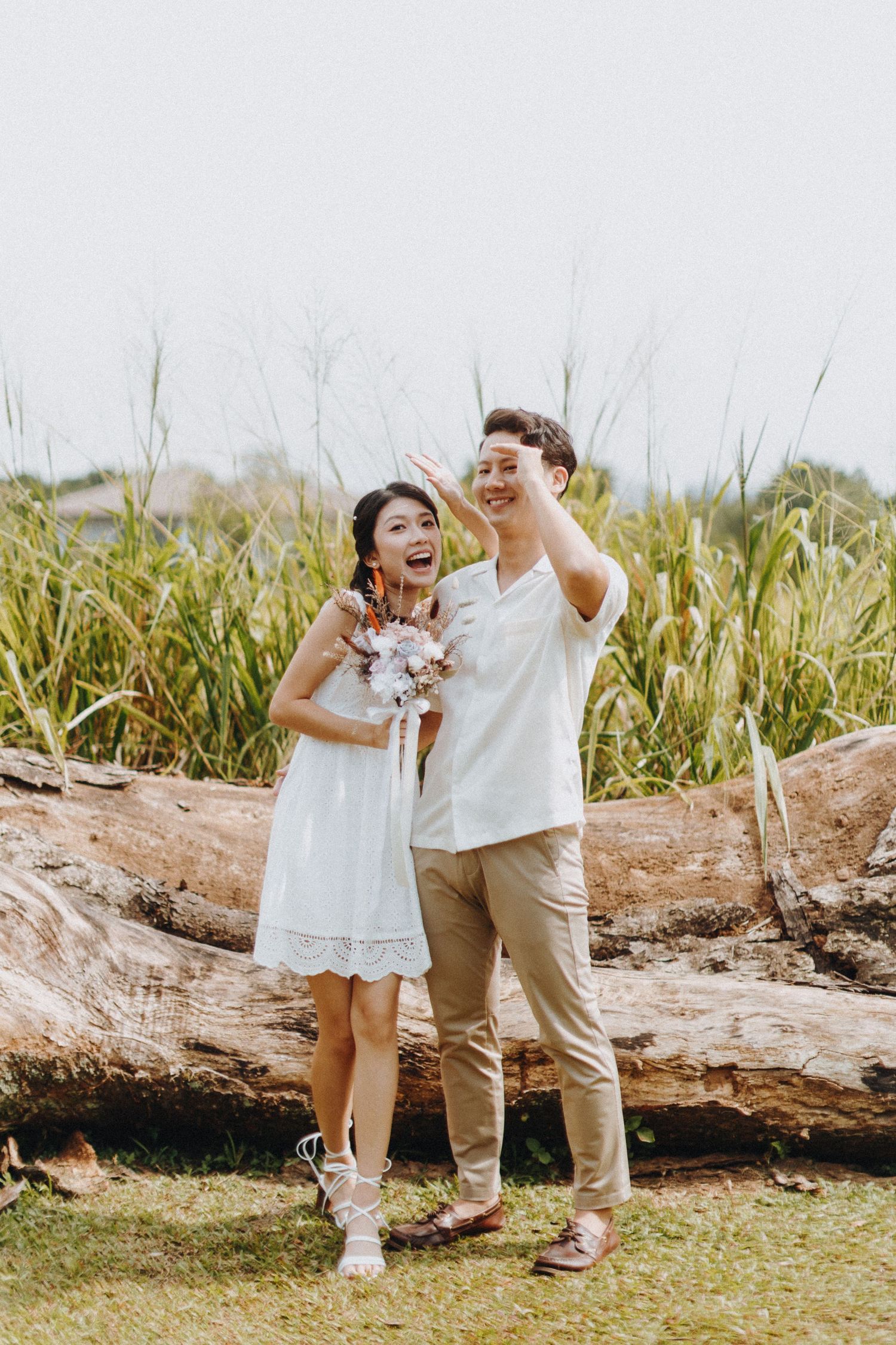 A couple in white outfits laughs together while standing on a fallen log surrounded by tall grass.
