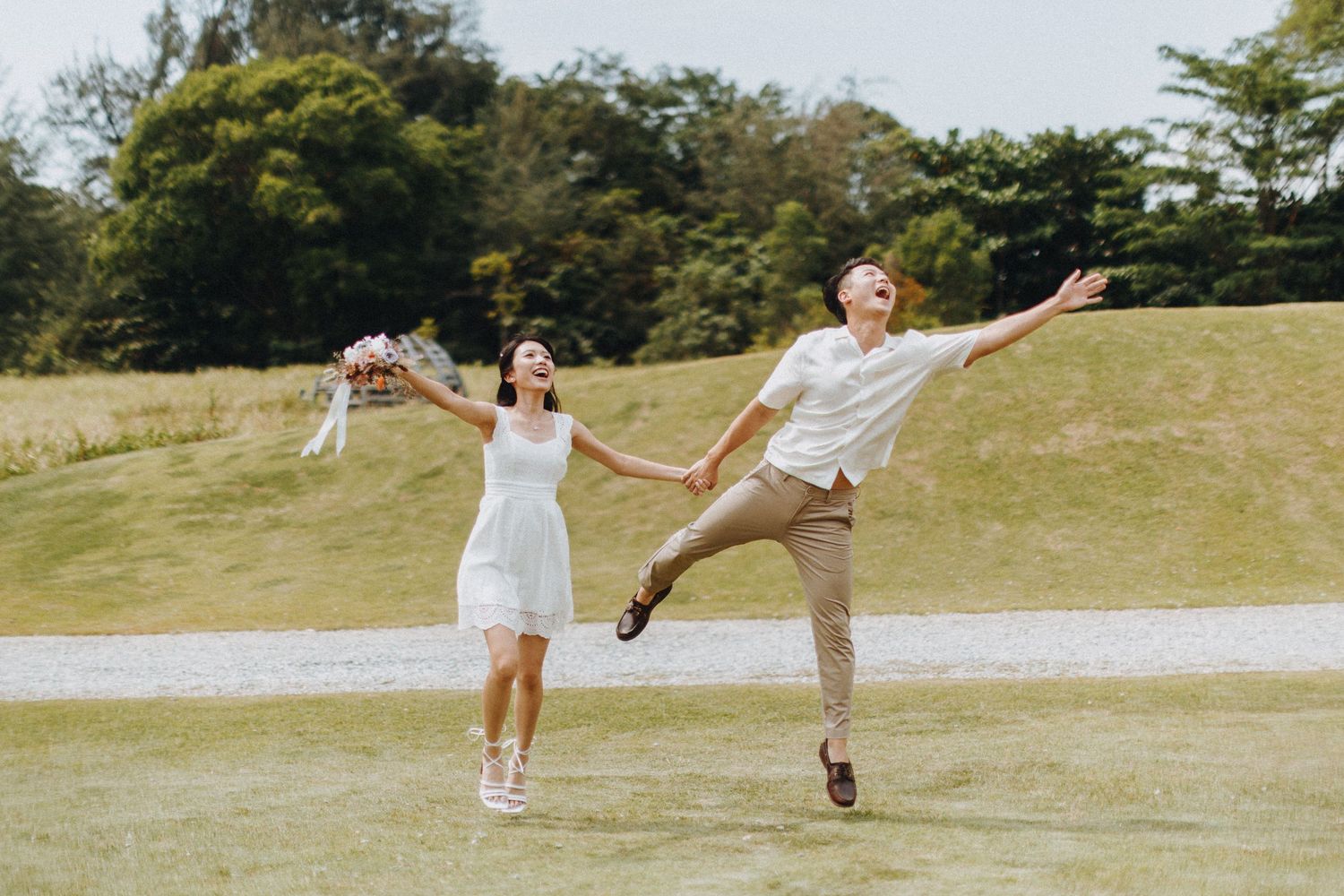 A couple in white and beige outfits jumps playfully together on a grassy hill.