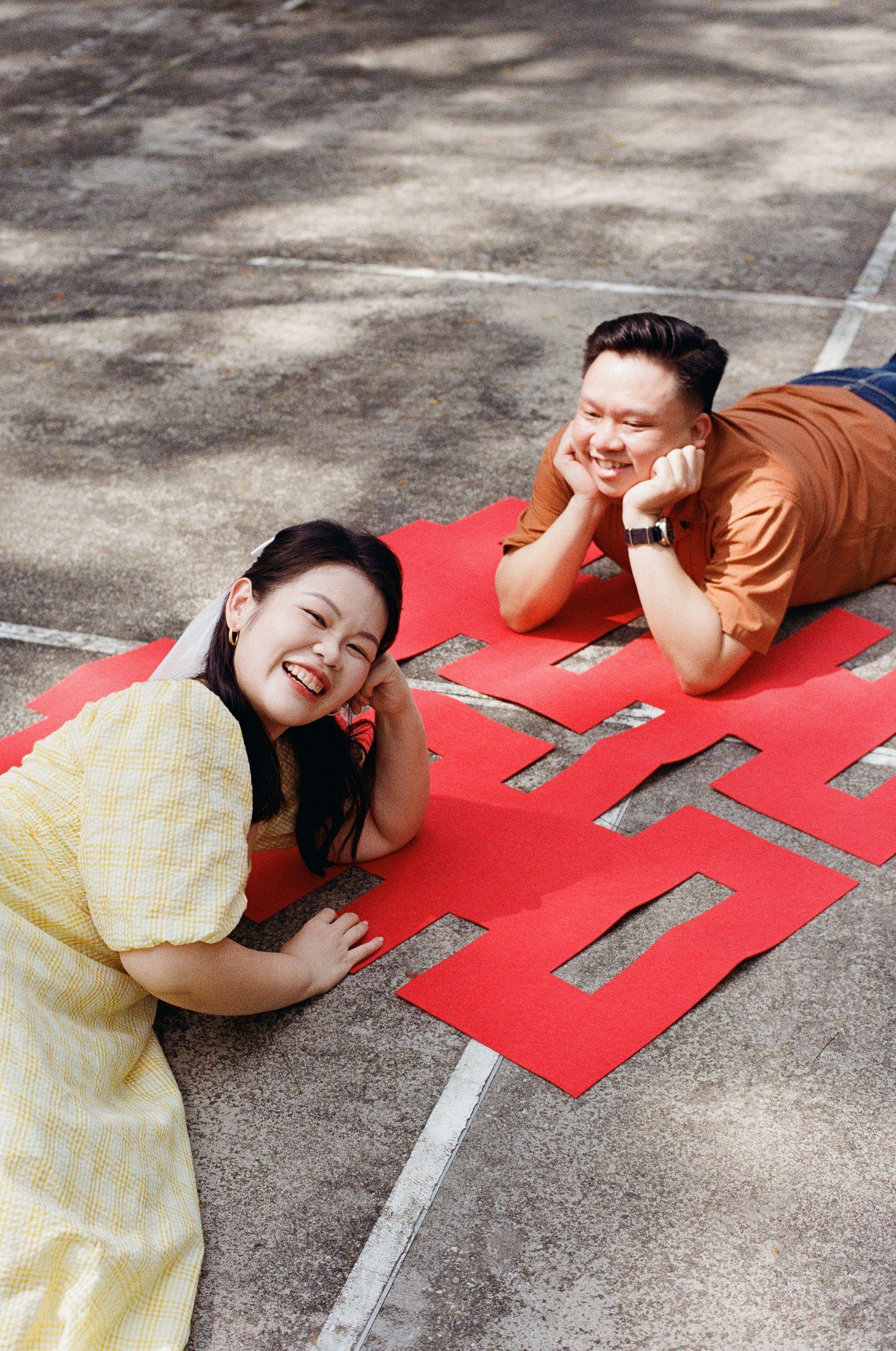A couple lies on a red paper decoration while posing playfully on concrete ground.