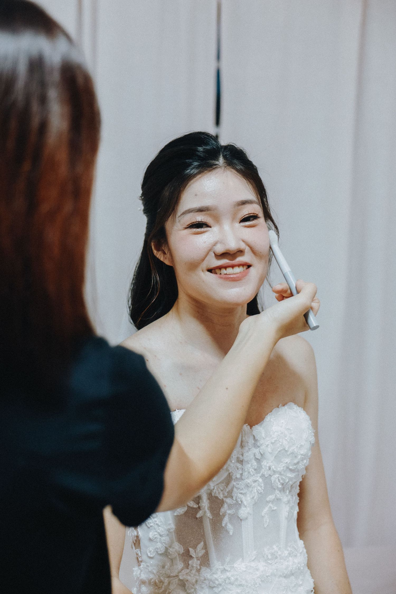 A bride having her makeup applied while wearing a white strapless lace wedding dress.