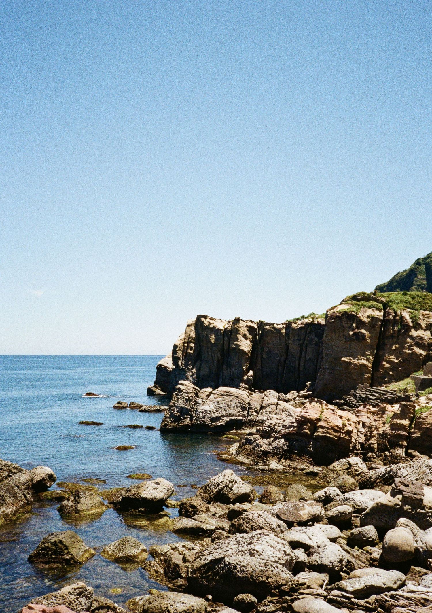 Rocky coastal cliffs rise above the blue ocean waters with scattered boulders along the rugged shoreline.