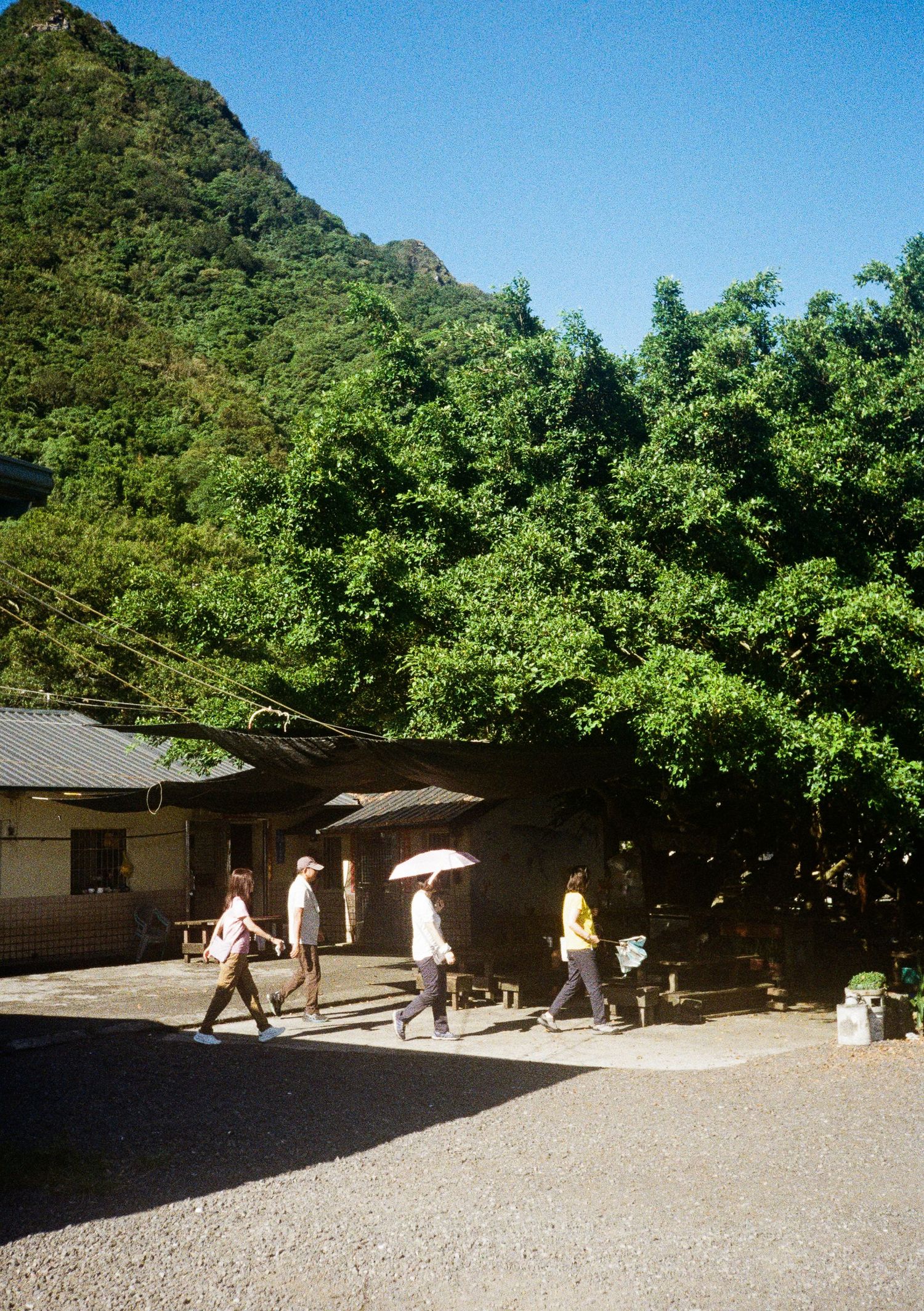 A series of rustic buildings nestled against a lush mountainside with people walking along a gravel path.