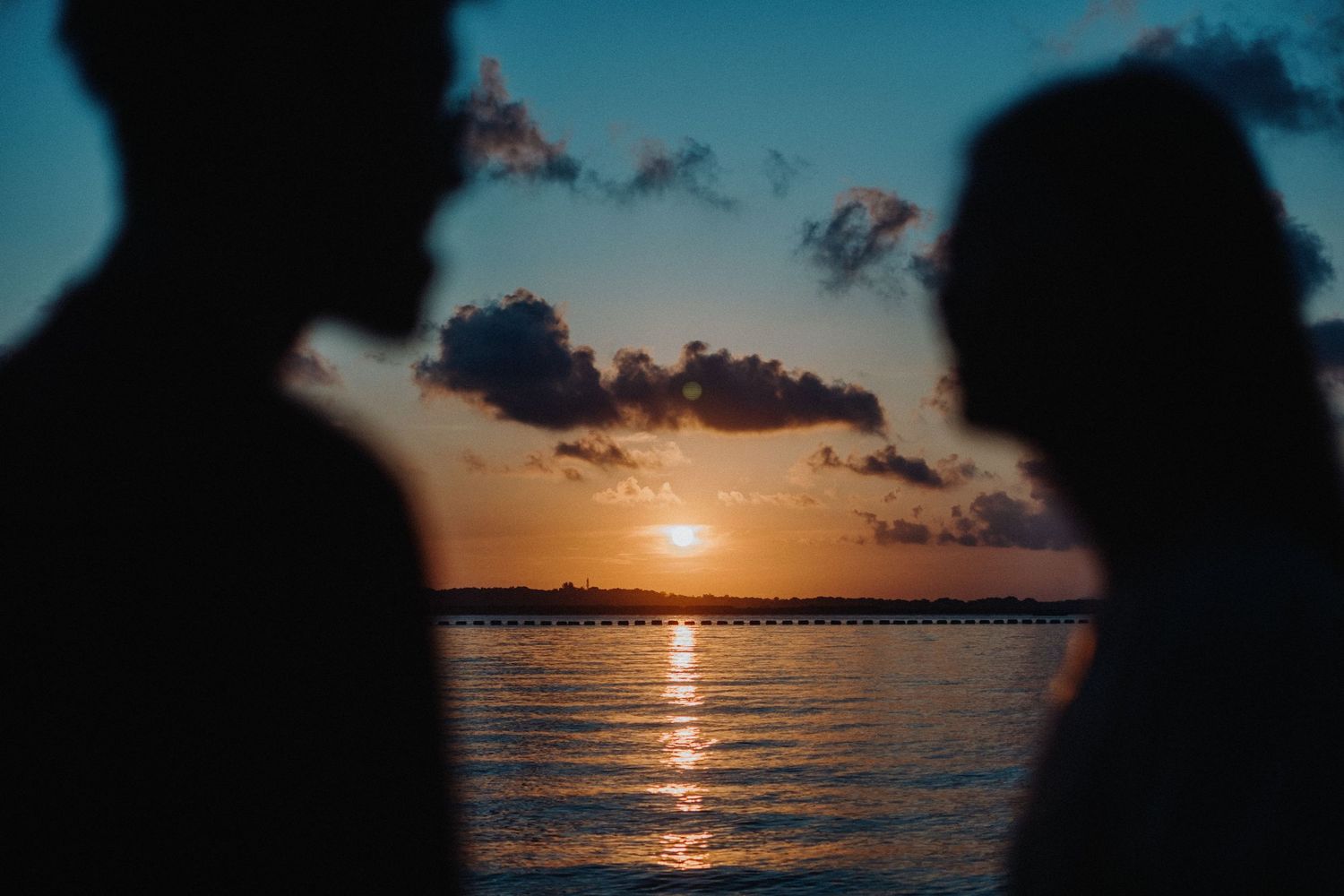 Dramatic silhouettes of people against a vibrant orange and blue sunset sky over calm ocean waters.