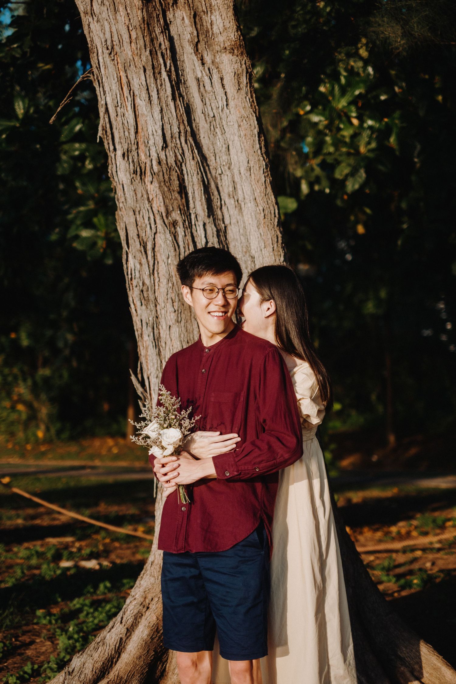 A romantic couple embraces by a large tree trunk in a park setting, with one holding a small flower bouquet.