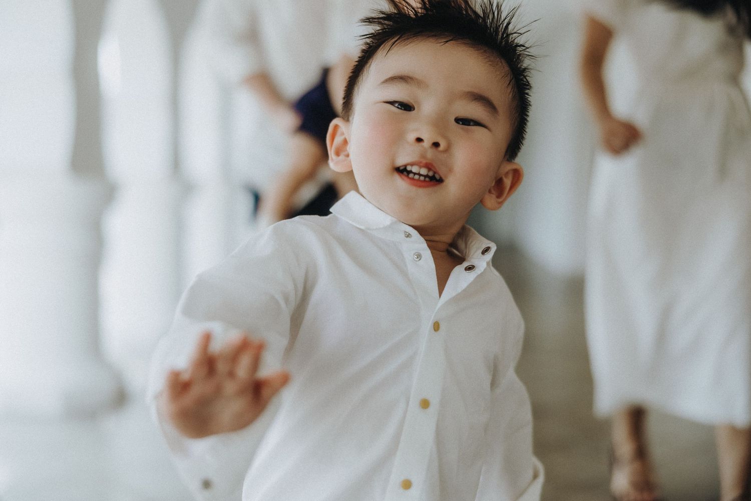 A young child in a white button-down shirt lies on white bedding while playfully reaching toward the camera.