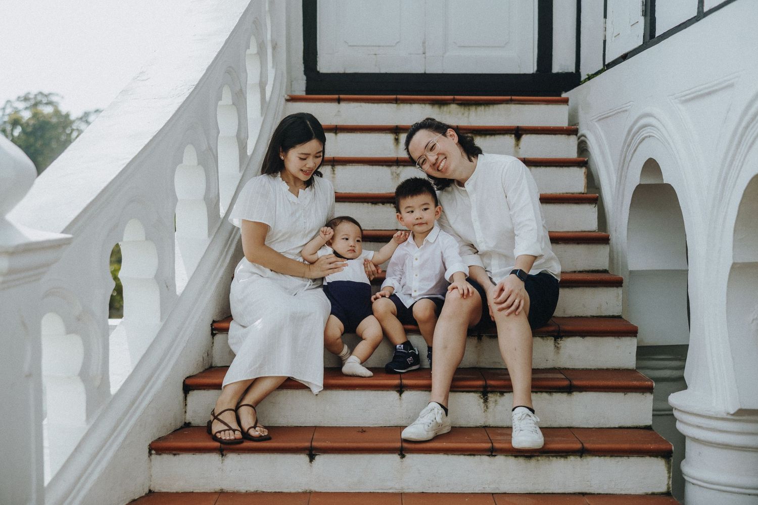 A family wearing white and cream colored outfits sits together on wooden stairs with white architectural details.