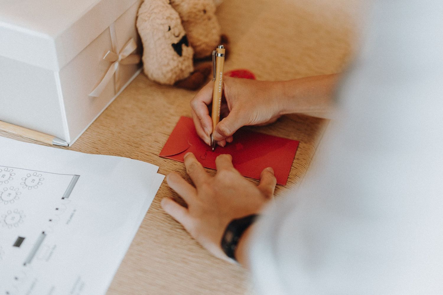 Hands write in notebook with red paper and scissors on wooden desk, viewed from above through white vignette.