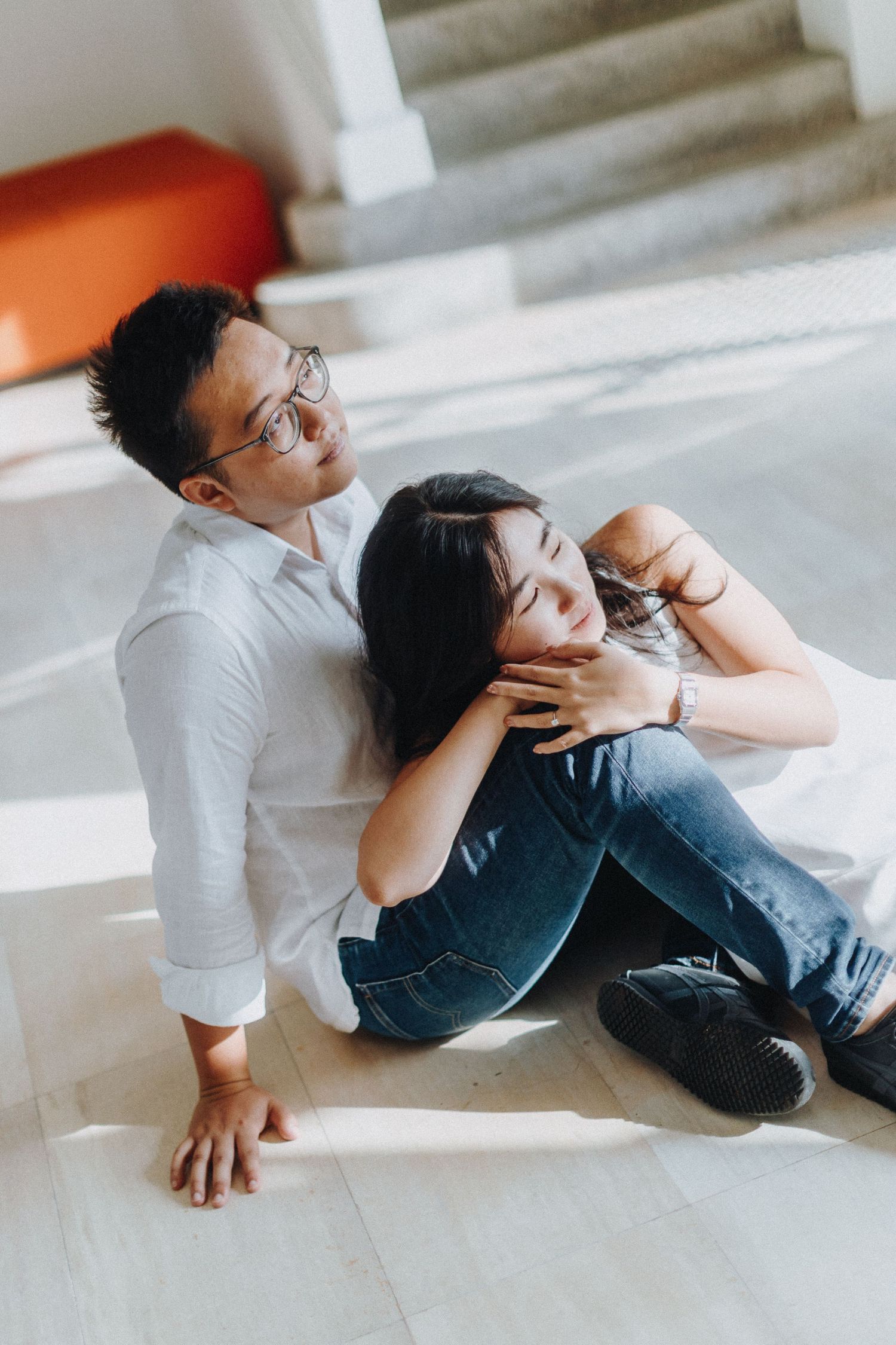 A couple relaxes on white steps in casual attire during an indoor photoshoot.