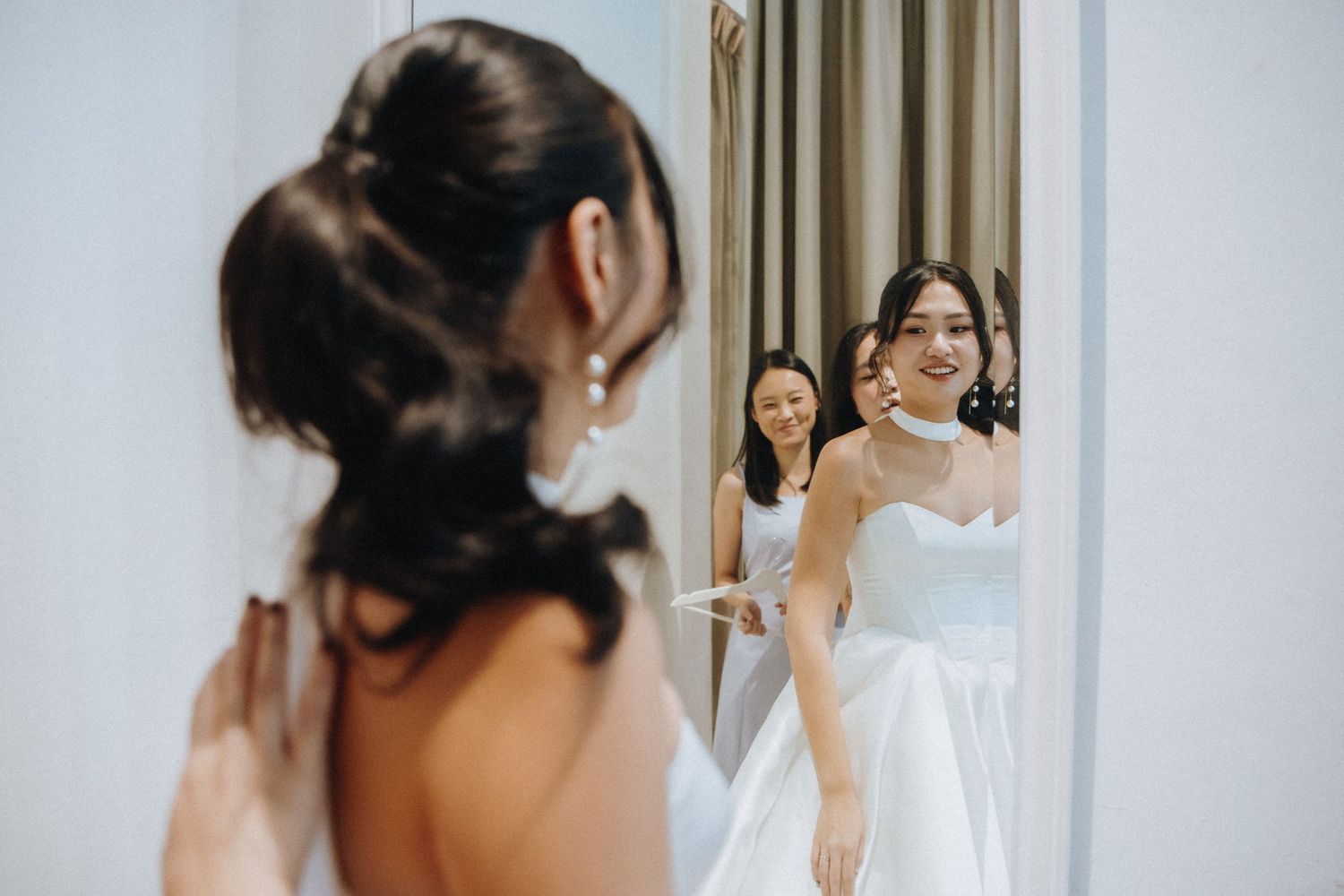 Bride in white wedding gown admiring her reflection in mirror with bridesmaid assisting her.