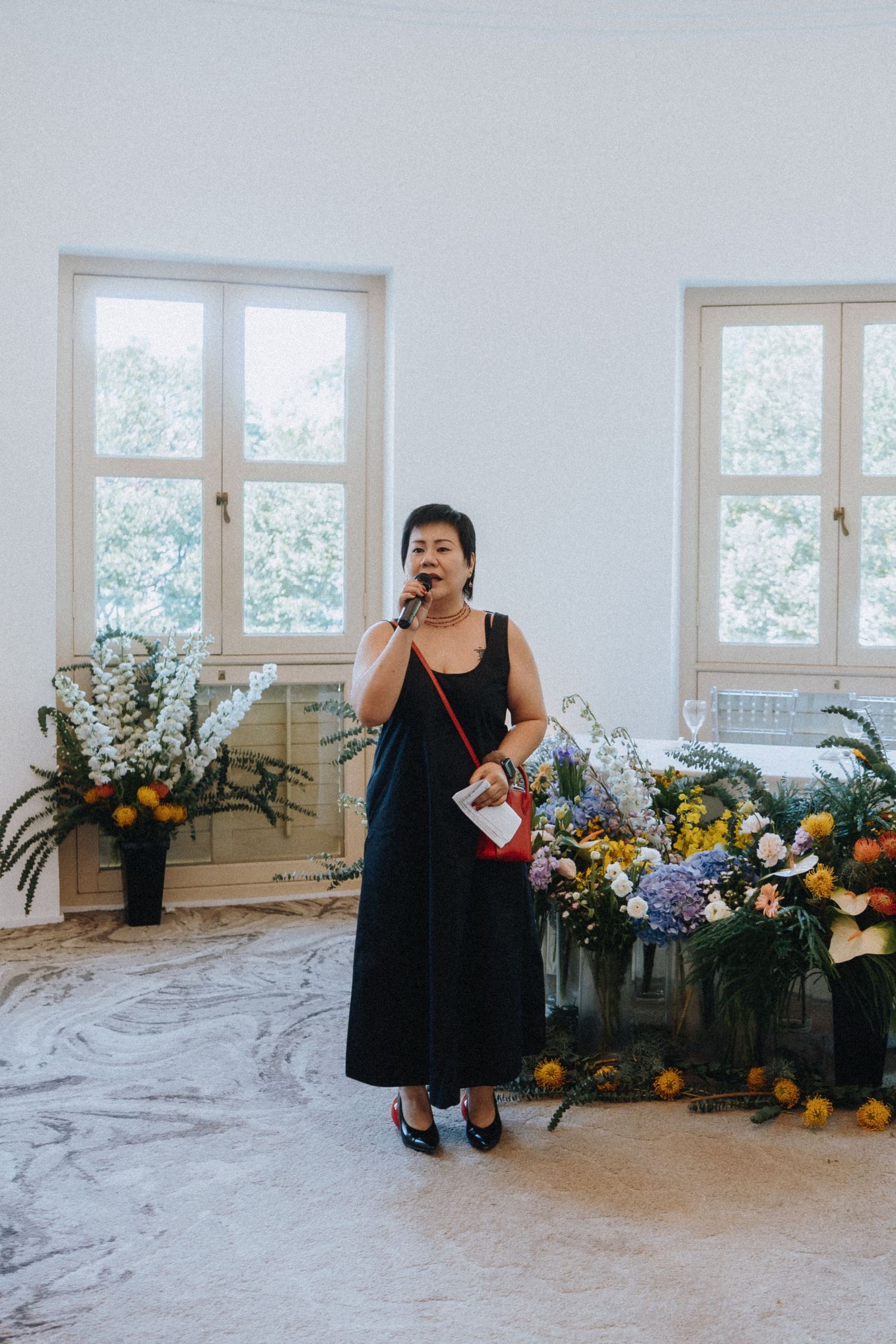 Woman in black dress speaks into microphone at elegant floral wedding ceremony venue.