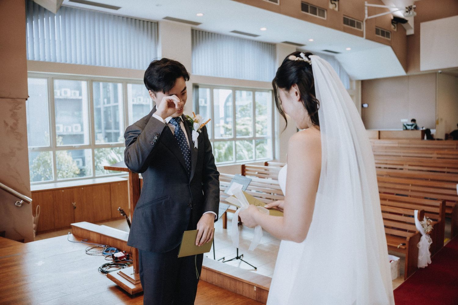 Groom wiping tears during emotional wedding vow exchange with bride in white dress inside a church.
