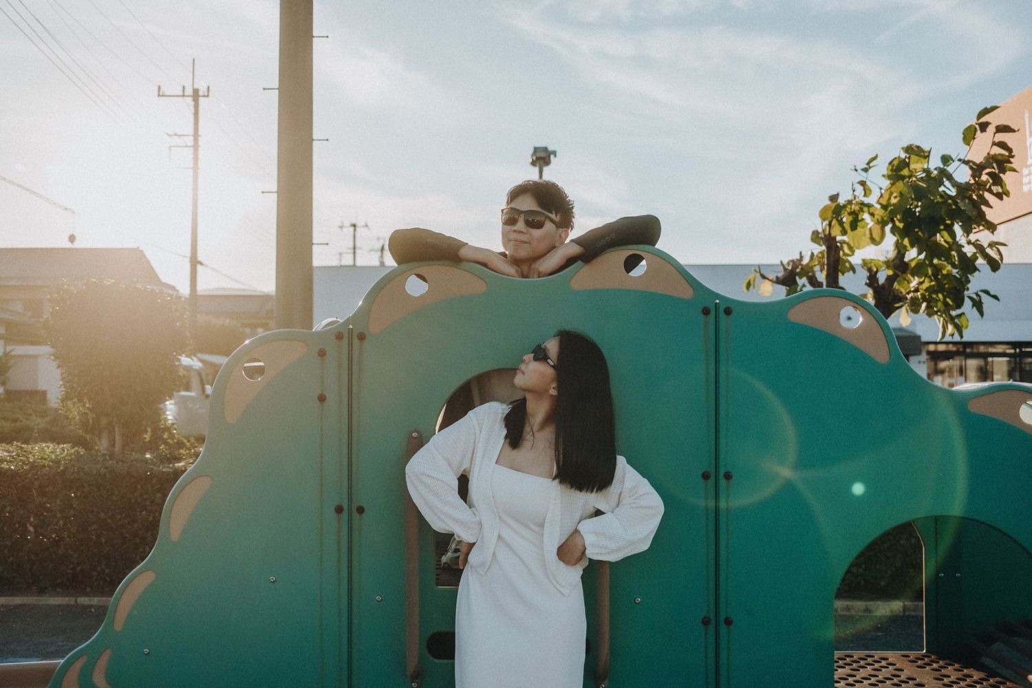 Playful couple in casual clothes posing against green storage containers at sunset.