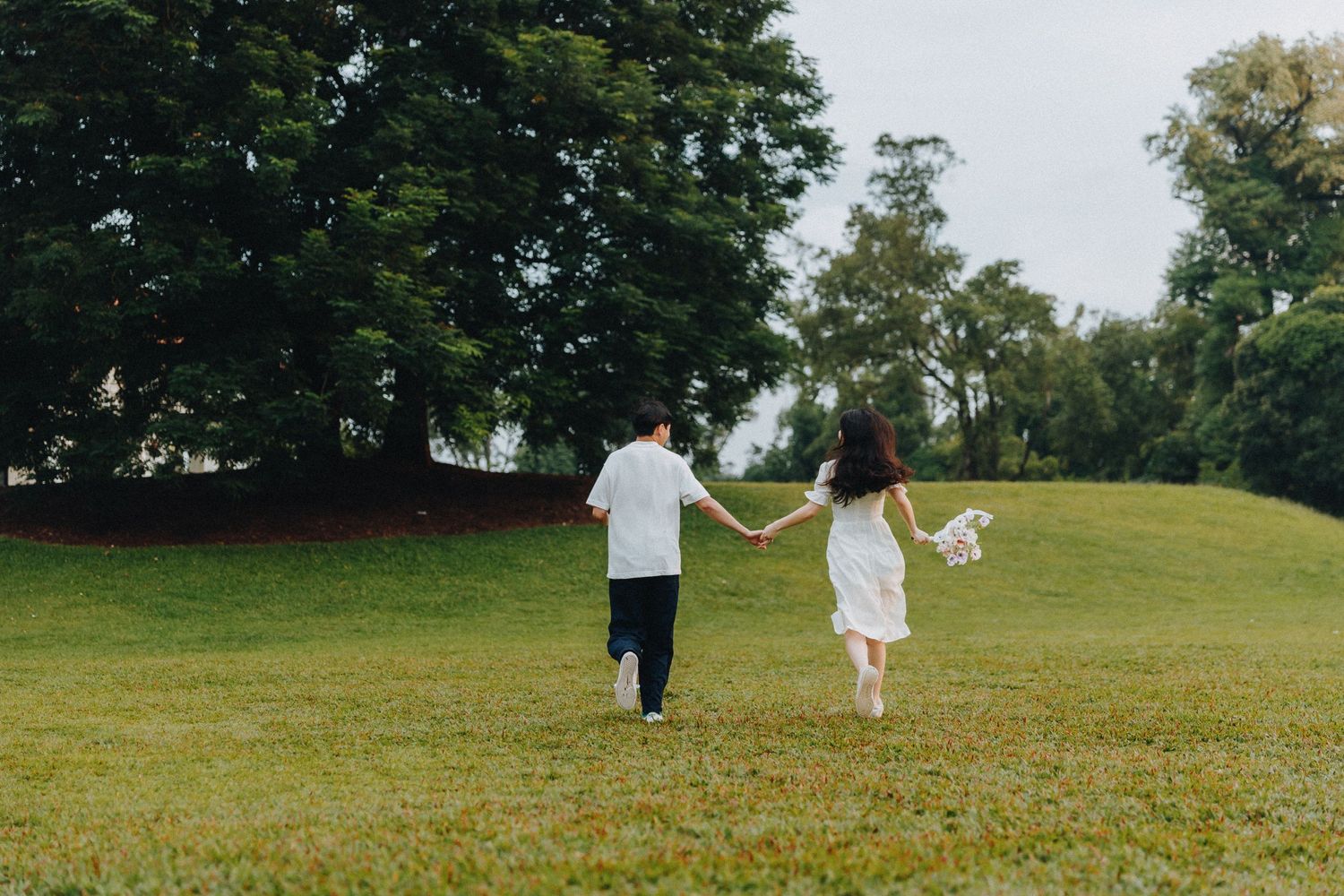 A couple holds hands while walking together across a grassy lawn surrounded by trees on a summer day.