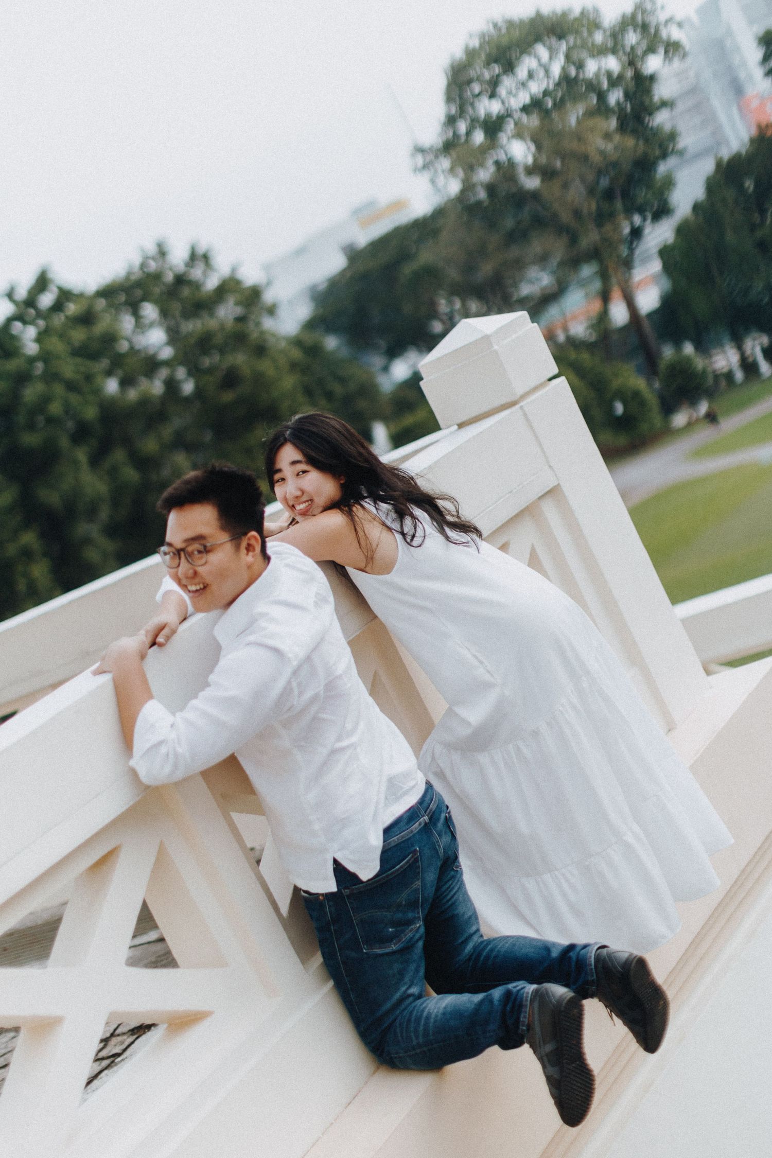 A playful couple in white clothing pose on a white railing outdoors.