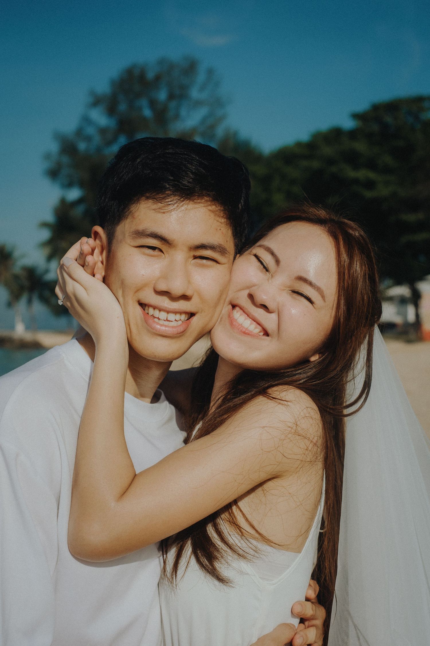 A joyful couple in white tops share intimate moments while laughing and embracing during a sunny outdoor photoshoot.