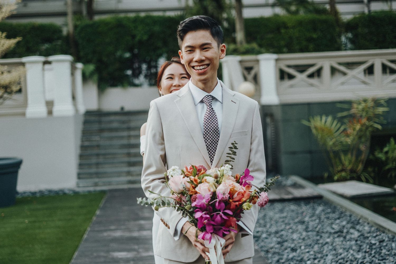 Wedding guest in a cream-colored suit holds a vibrant pink and purple floral bouquet while standing outdoors.