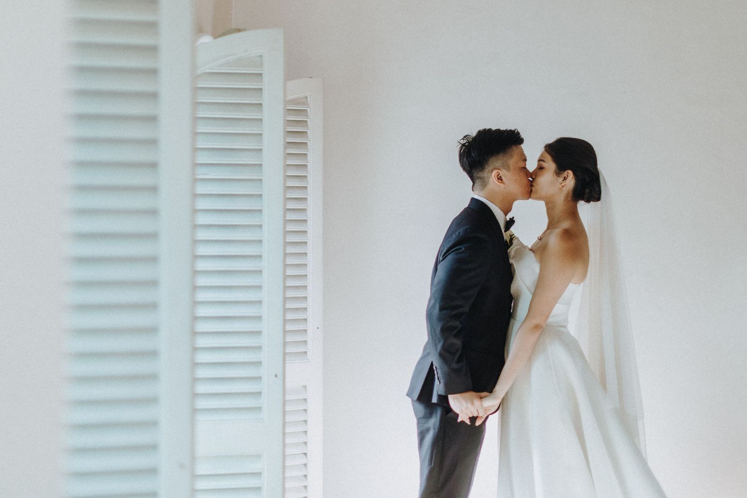 A romantic wedding scene with a couple sharing a kiss next to white shutters in a minimalist setting.