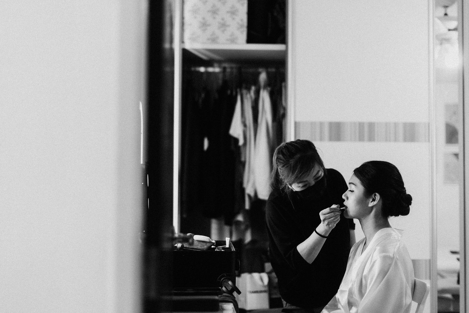 Black and white photo showing backstage preparations with someone having makeup applied while wearing a white robe.