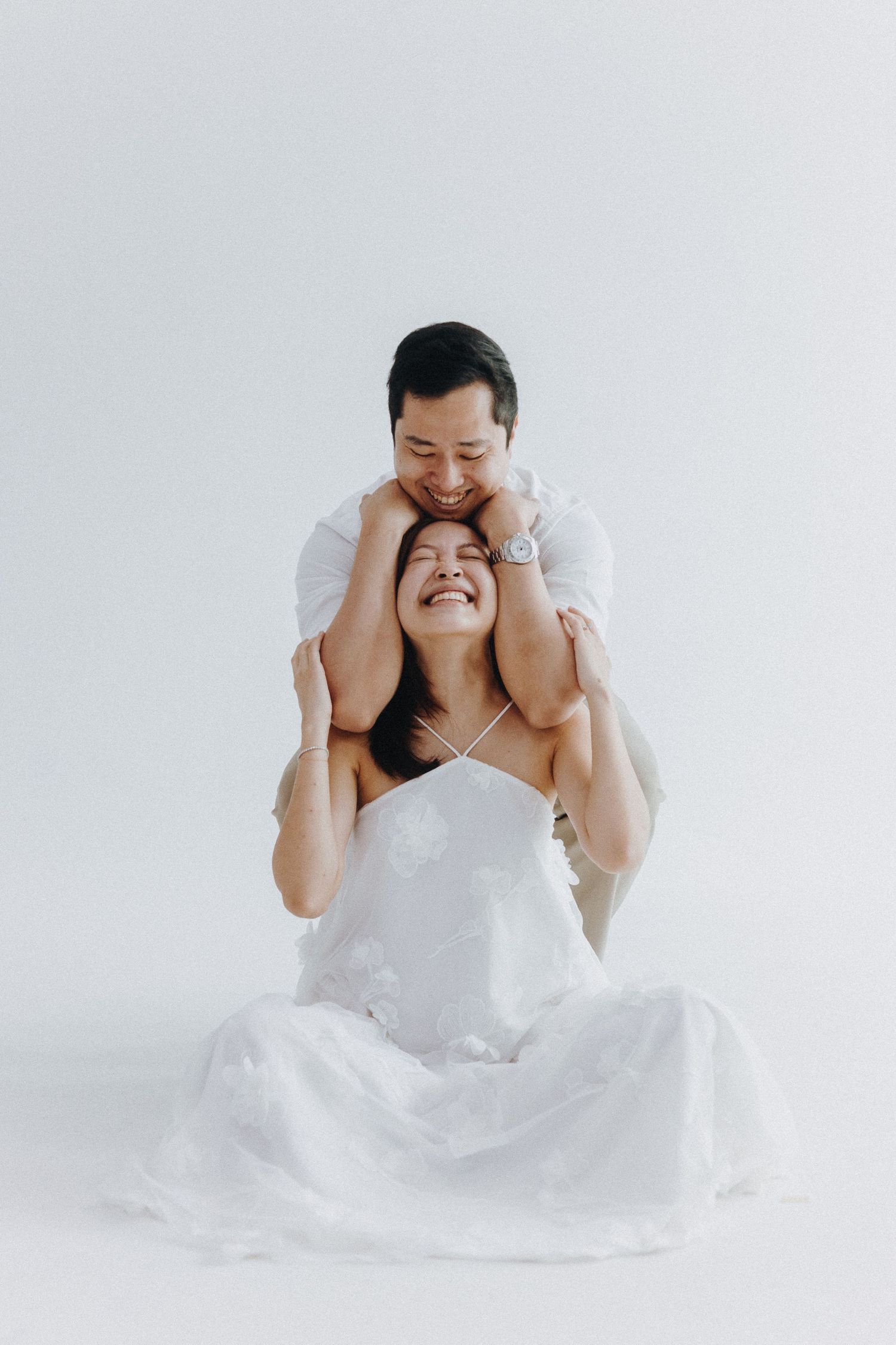 A playful couple in white attire sharing a joyous moment against a minimalist background.