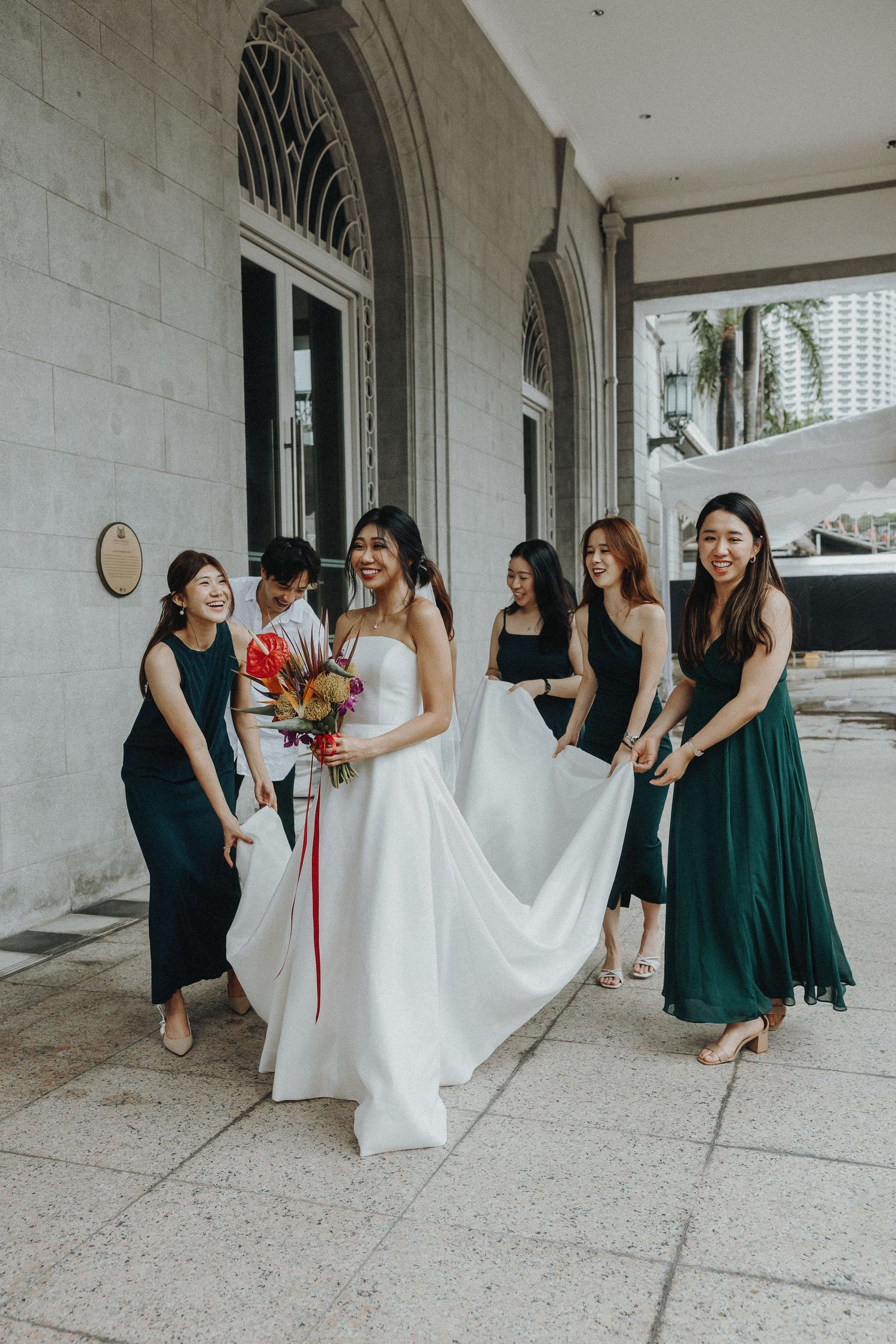 Bridal party with women in dark dresses helping arrange bride's white wedding gown on stone porch.