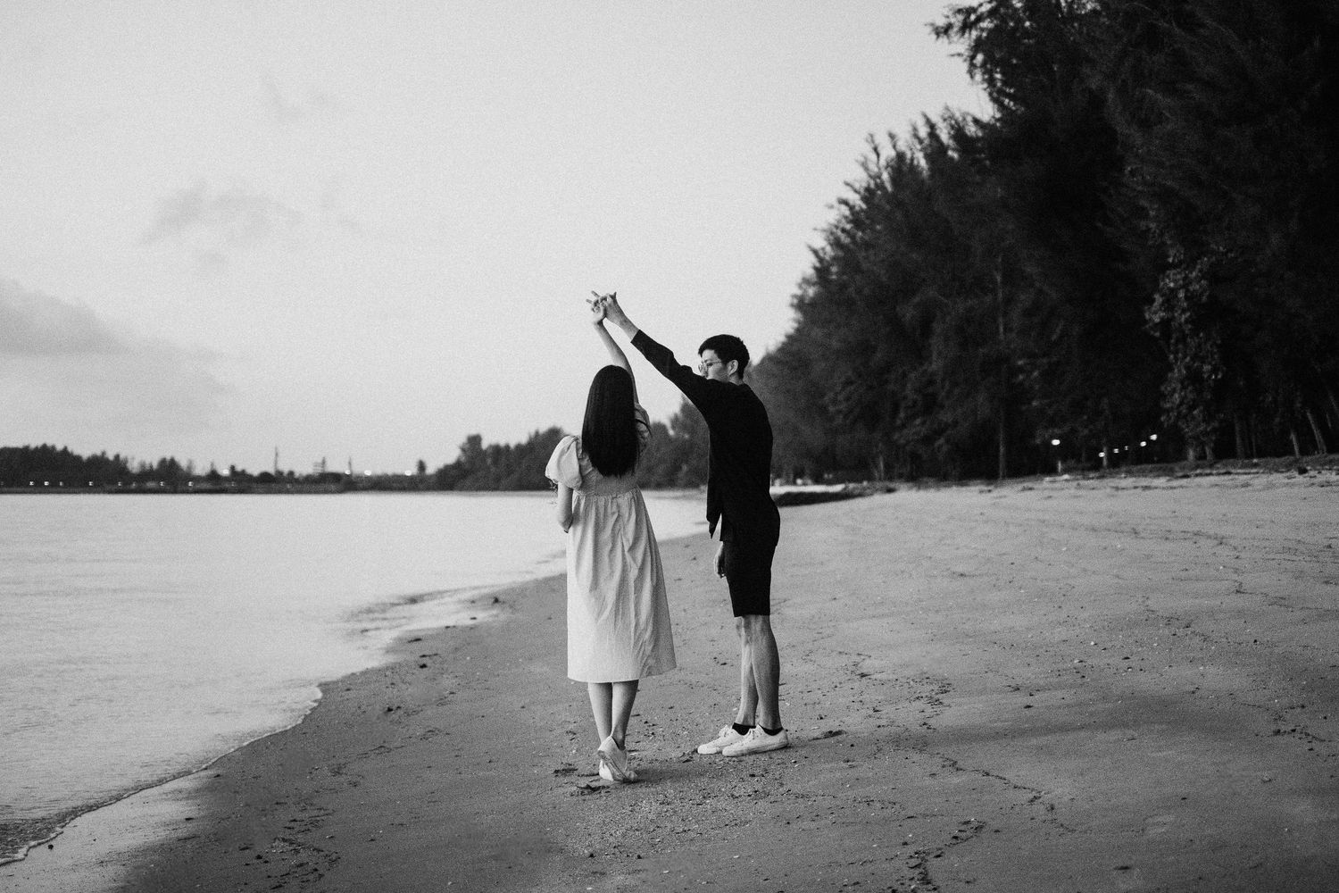 A couple in silhouette holds hands with raised arms on a serene beach with forest backdrop, captured in black and white.