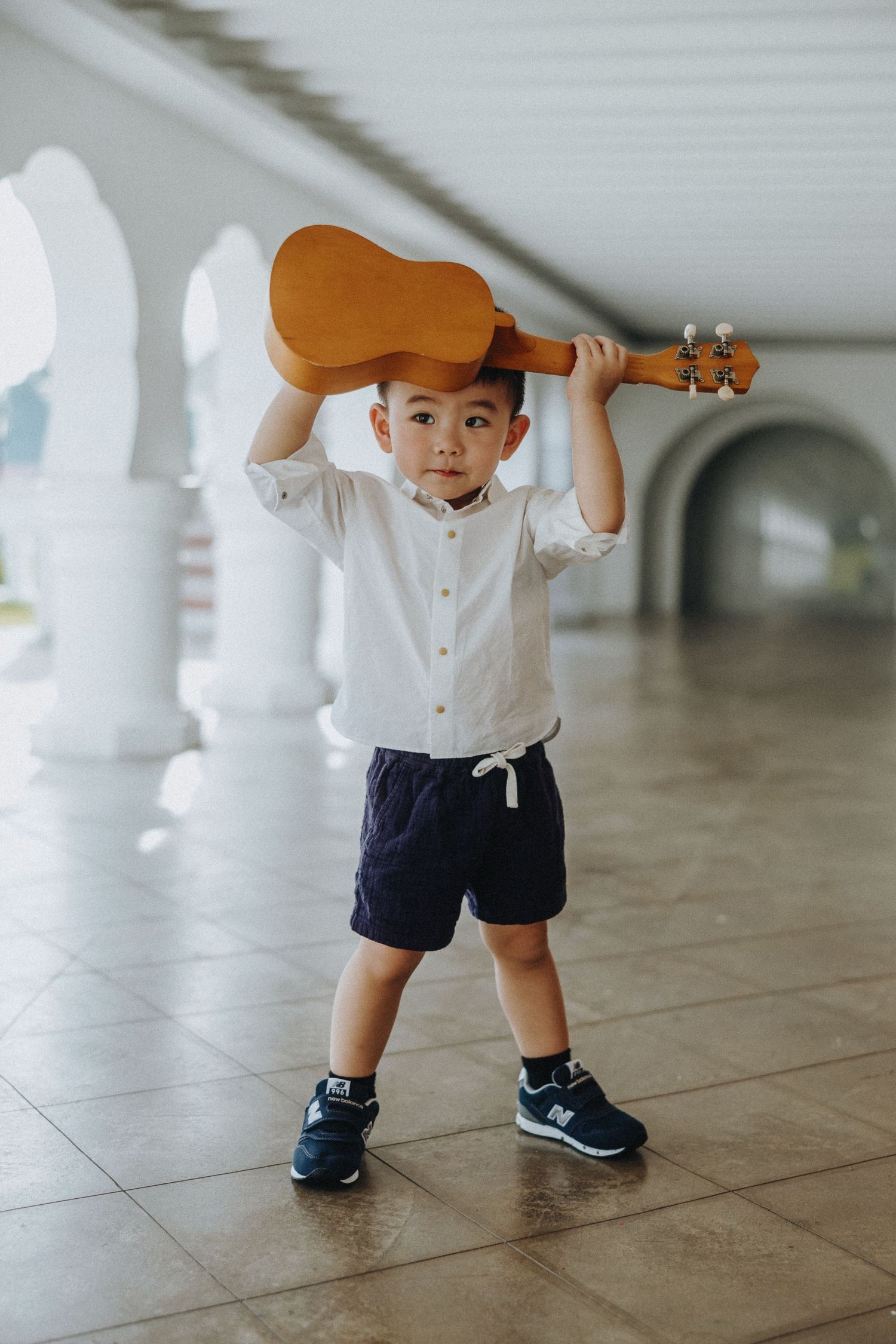 Young child in white shirt and navy shorts balances a ukulele on their head in a corridor.