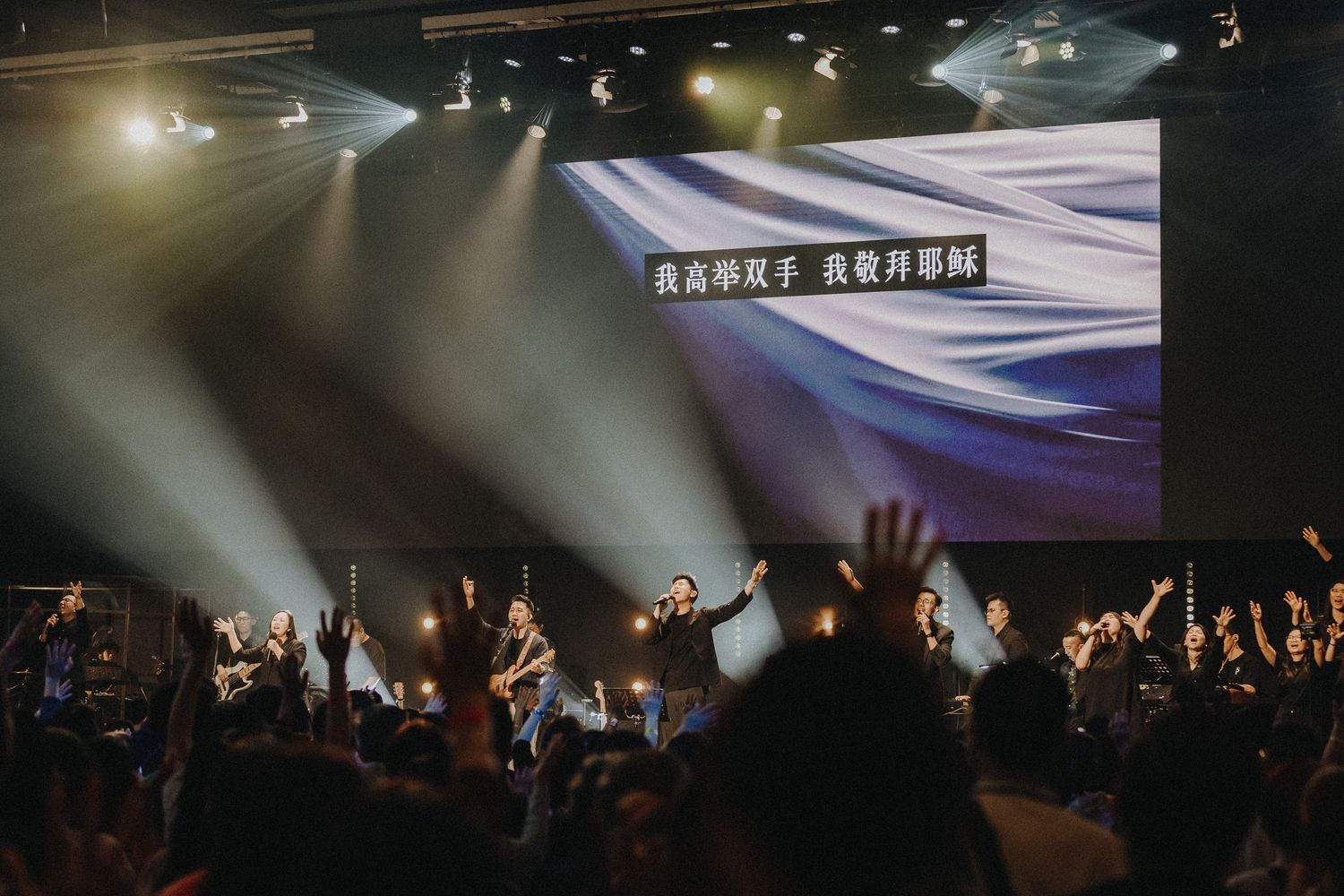 Silhouetted crowd with raised arms at concert venue with dramatic stage lighting and projection screen display.