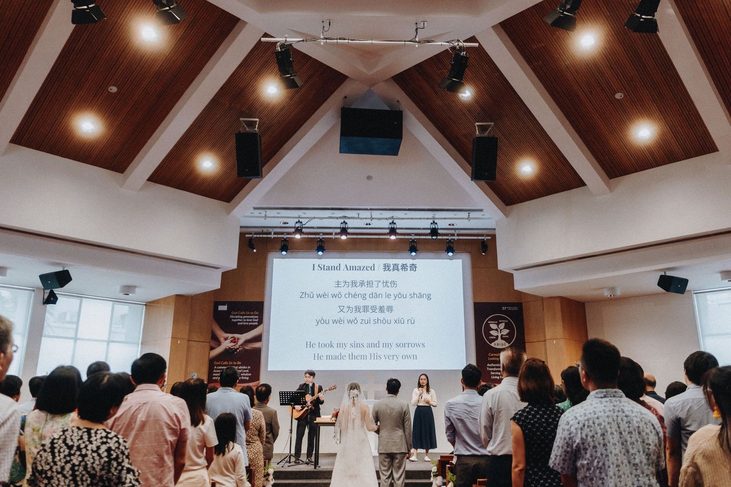 Church ceremony in progress with audience seated in rows under angled wooden ceiling with spotlights.