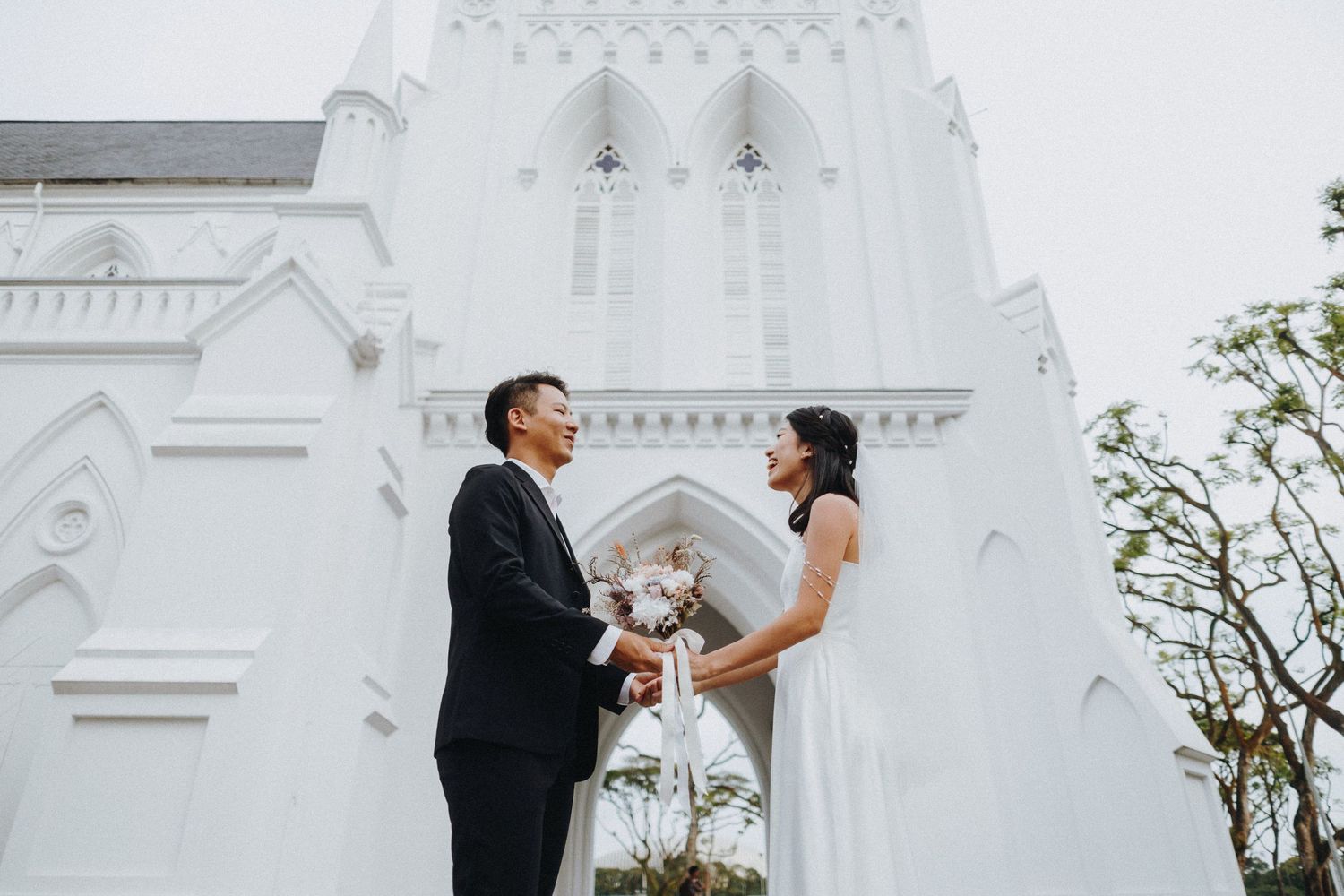 A couple poses in wedding attire in front of a white gothic church spire.