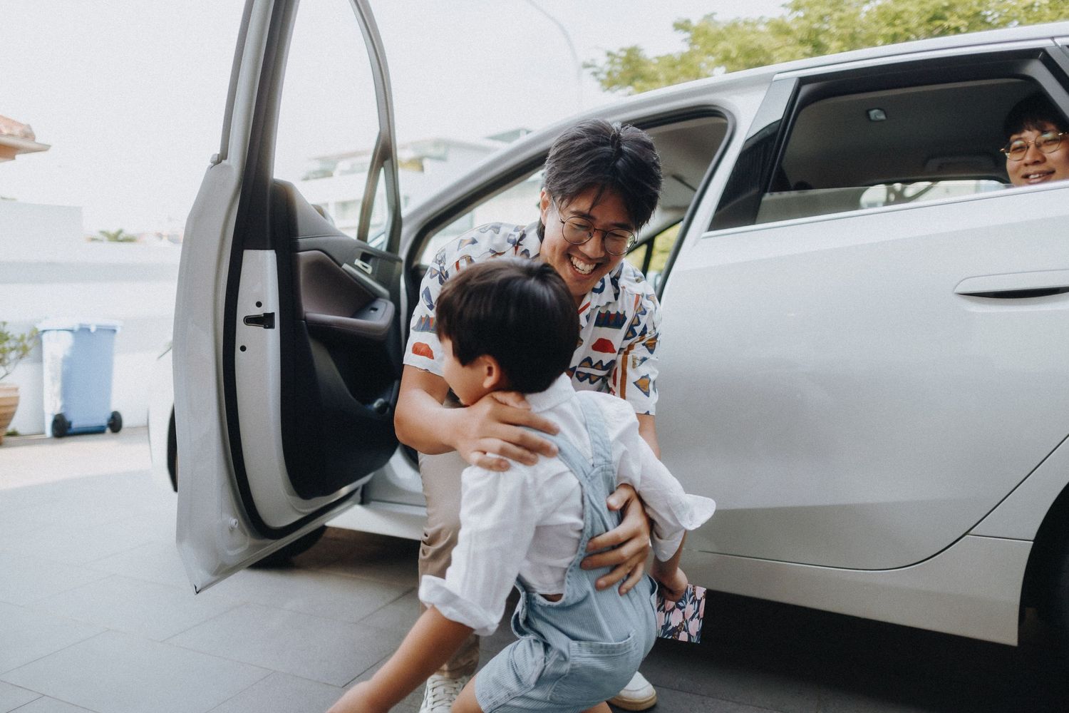A person helps a small child out of a silver car in a driveway on a sunny day.
