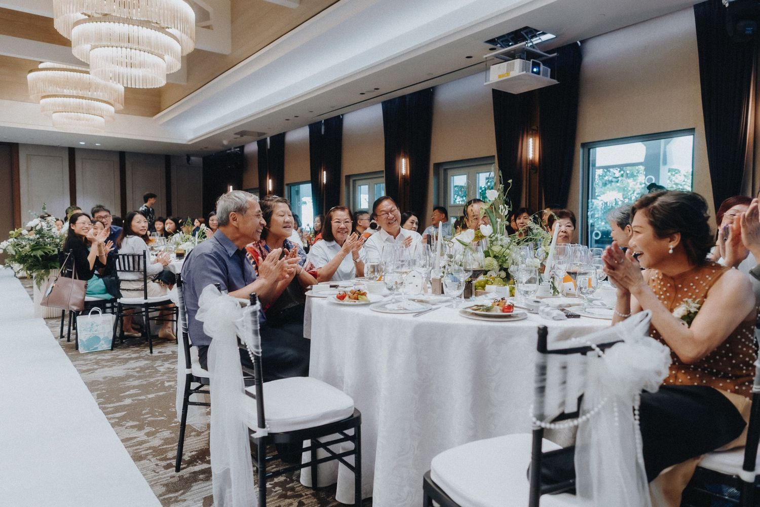 Wedding reception in a grand ballroom with chandeliers, round tables, and guests applauding.