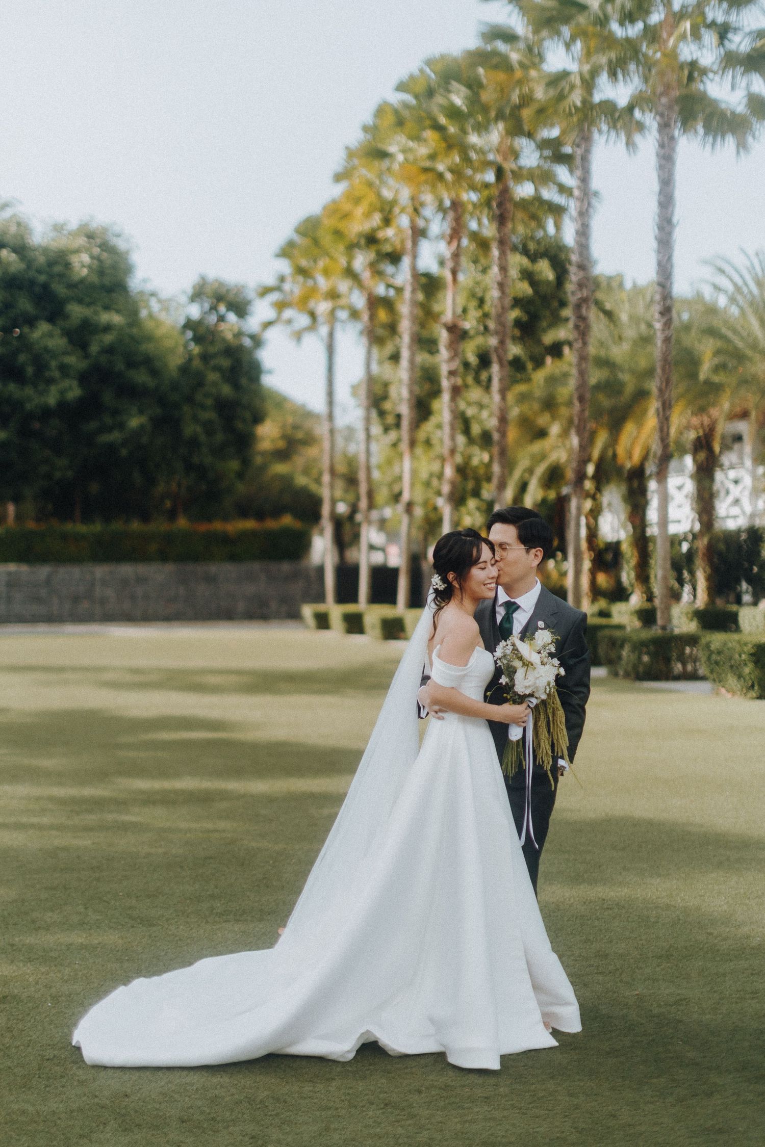 Newlyweds share a romantic moment on a manicured lawn surrounded by tall palm trees.