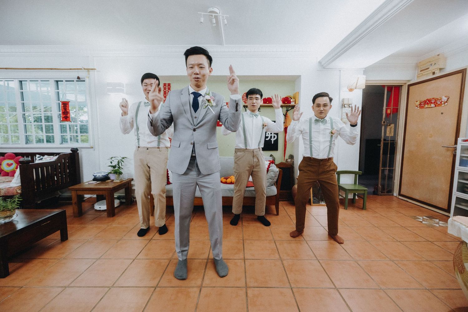 Groomsmen in gray suits and suspenders pose energetically in a living room.