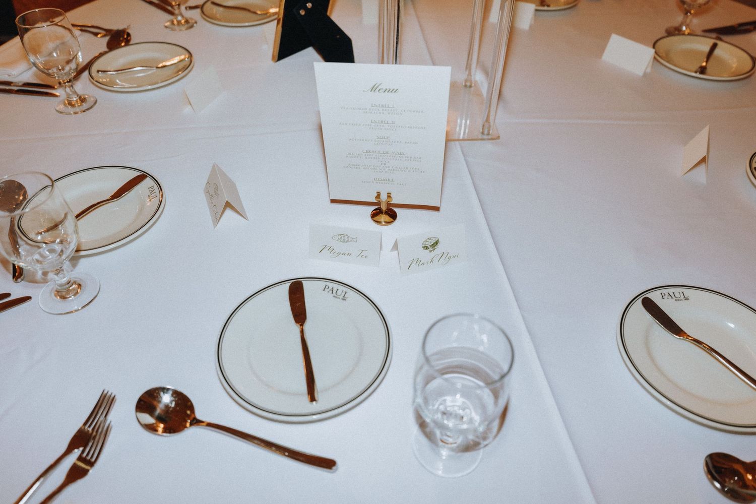 Elegant wedding reception table setting with gold cutlery, white plates, glassware, and a menu card on white linen.