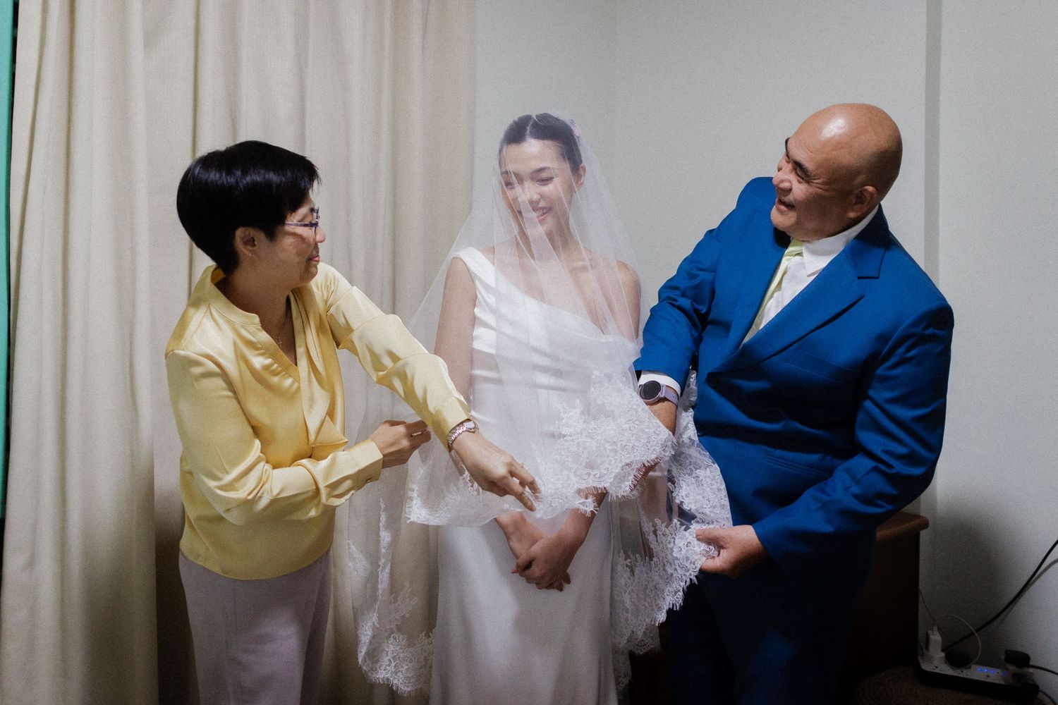 A bride in a white gown and veil stands between two smiling guests in a bright room during wedding preparations.