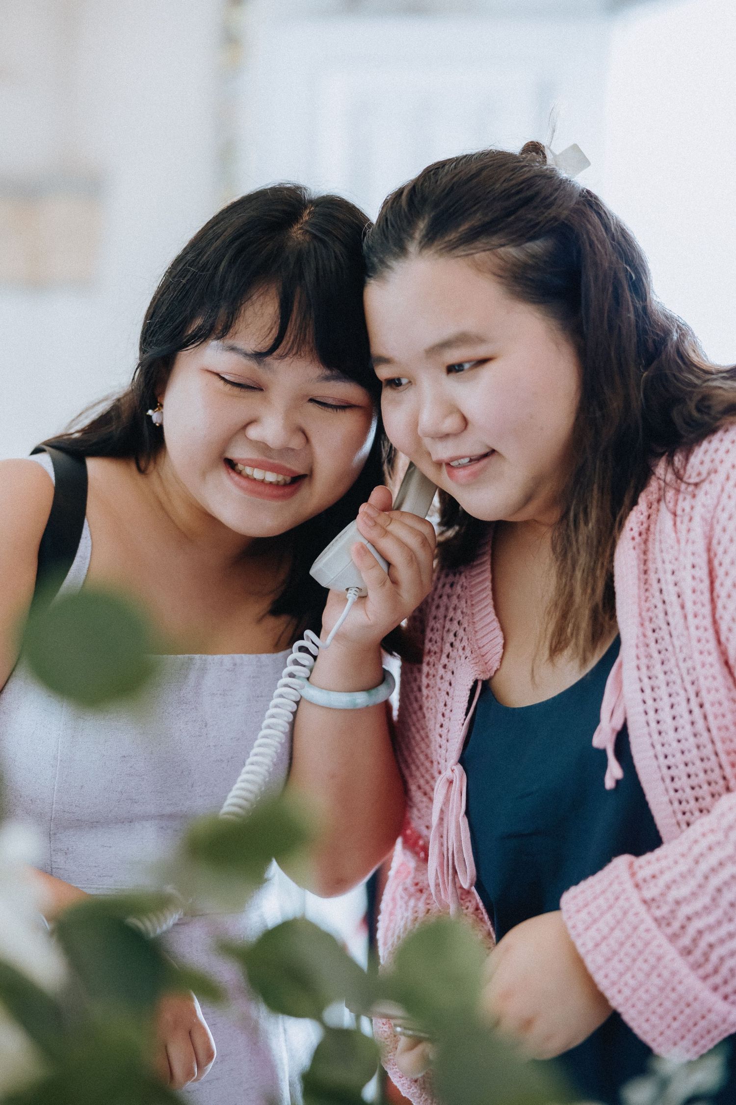Two young Asian women share a landline phone receiver, smiling and laughing together in a bright indoor setting.