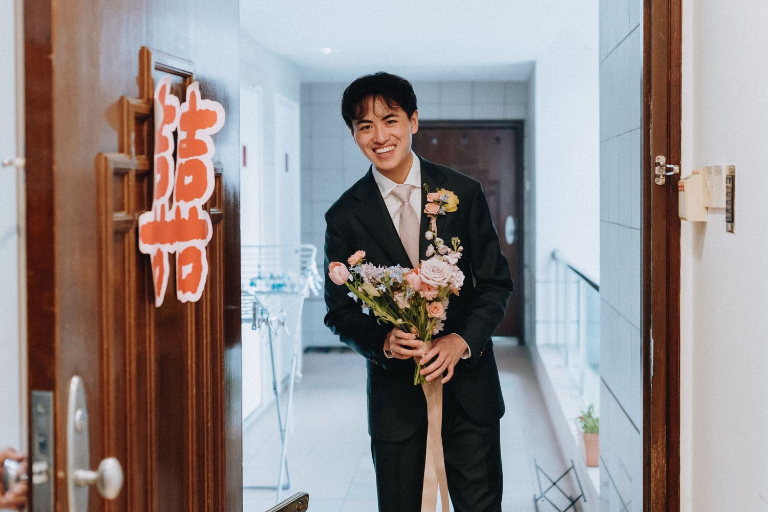 Smiling groom in black suit holding pink floral bouquet standing in doorway with red Chinese wedding character.