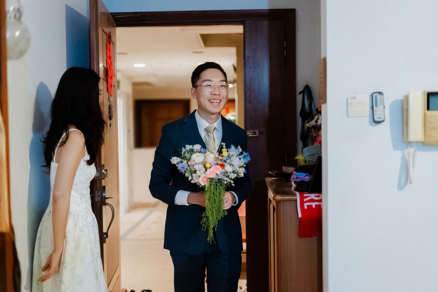 Groom in navy suit holding colorful bridal bouquet smiles at bride in white dress during wedding morning preparation.