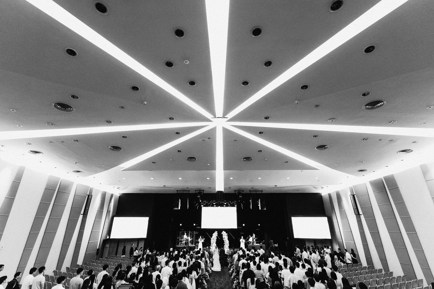 Black and white wide-angle shot of a large wedding ceremony hall filled with guests.