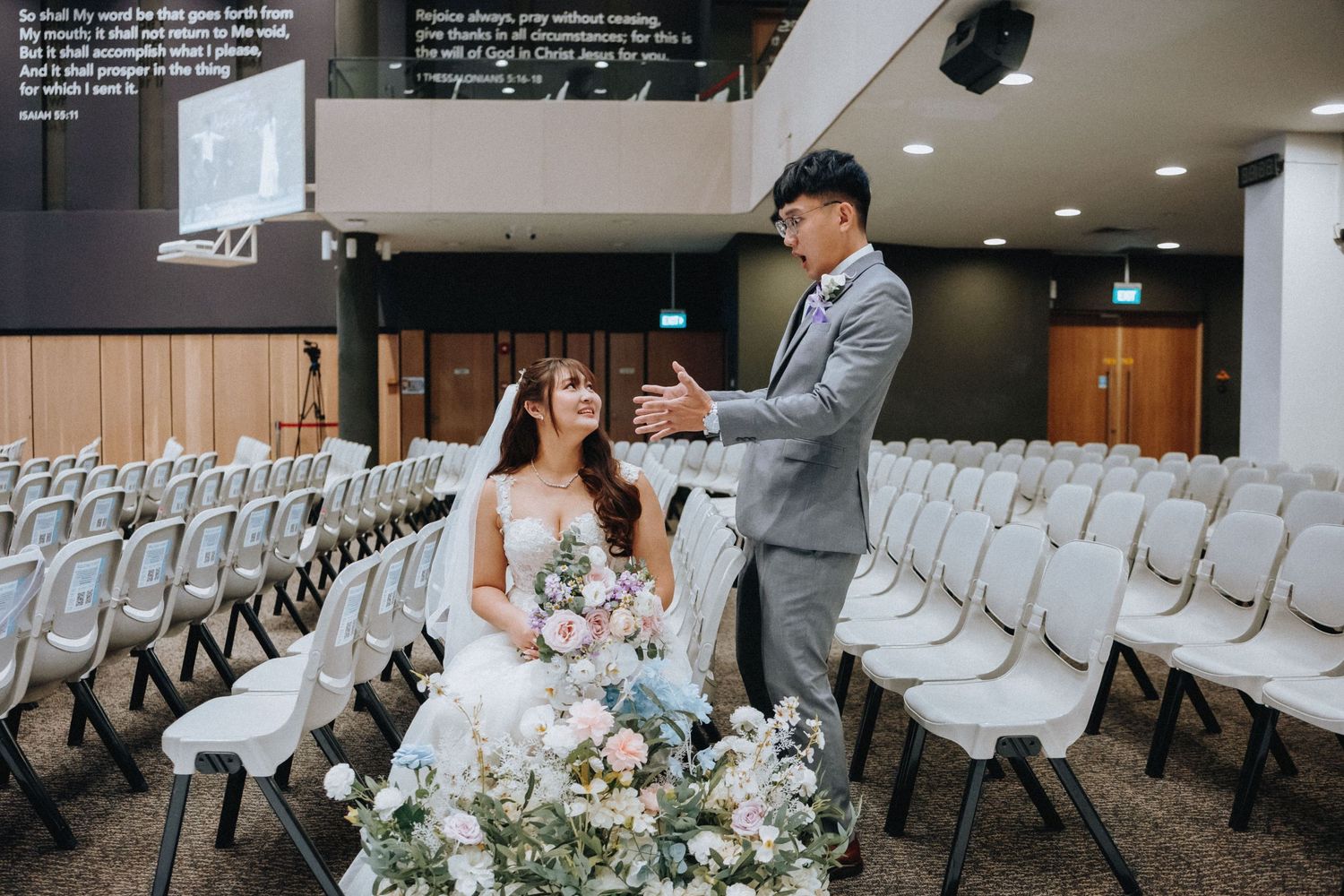 A wedding couple poses together in an auditorium with white chairs and floral arrangements.