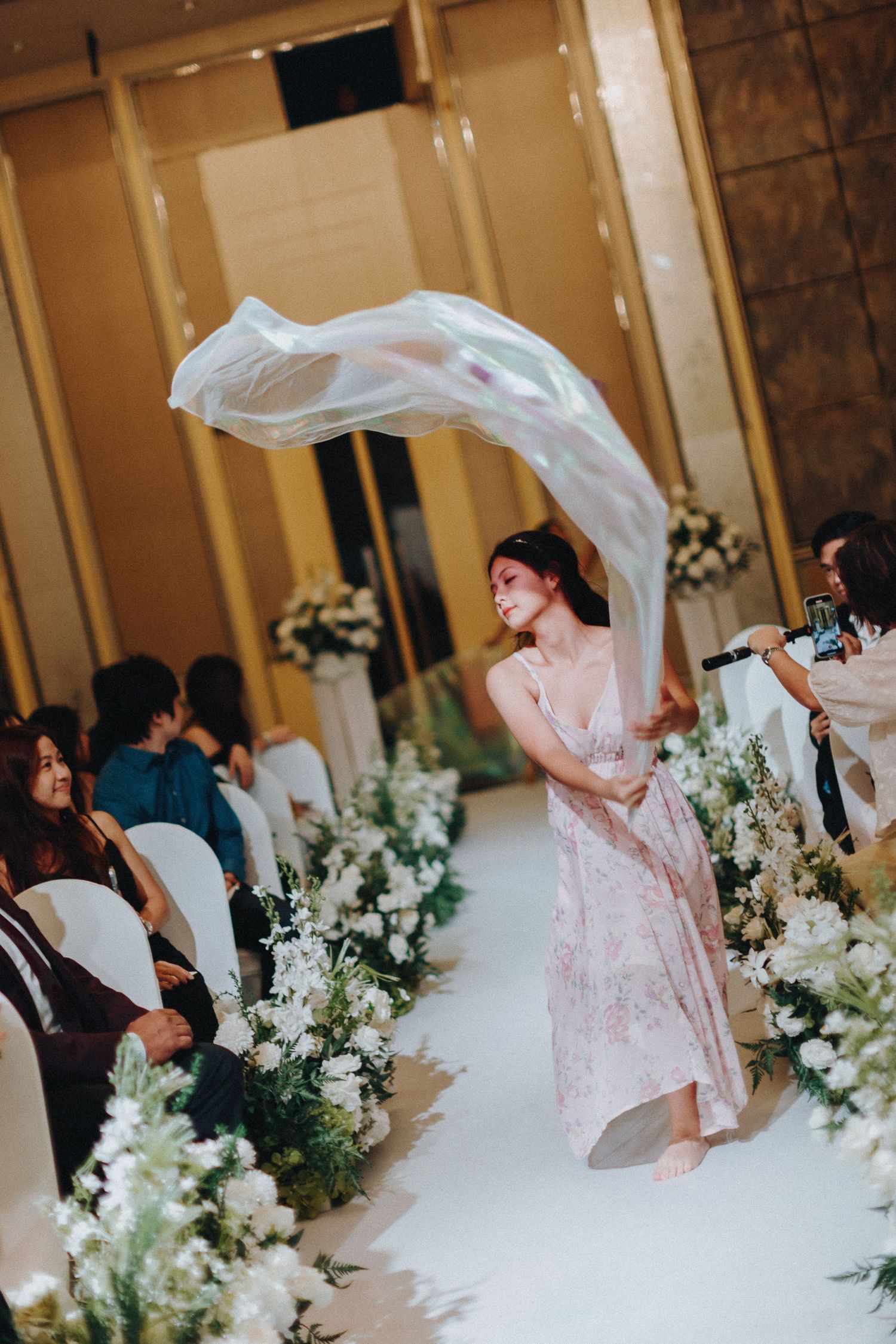 A person in a pink dress twirling a white veil during a wedding ceremony with floral decorations.