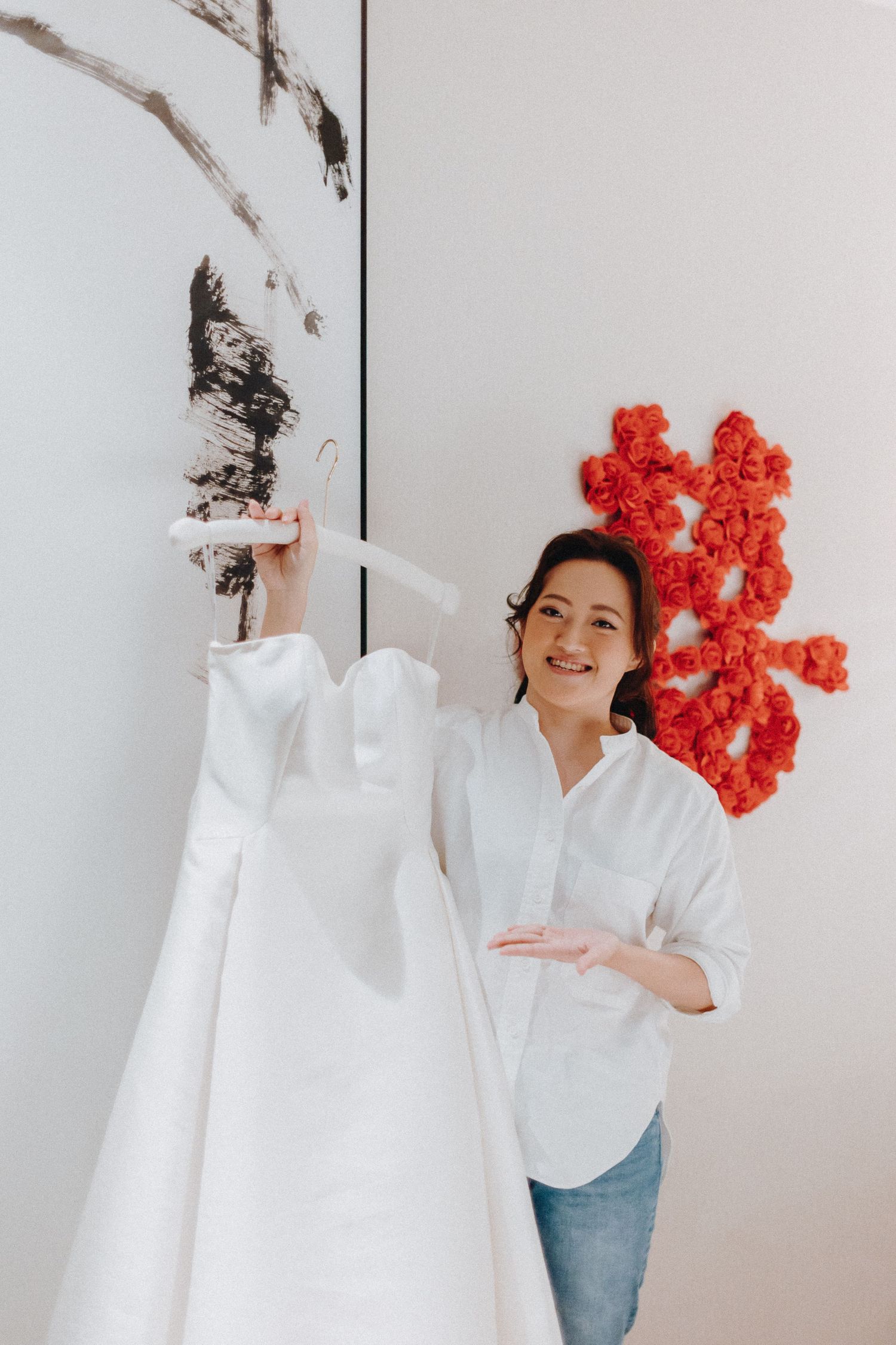 A white wedding dress is displayed against a wall with red floral decorations.