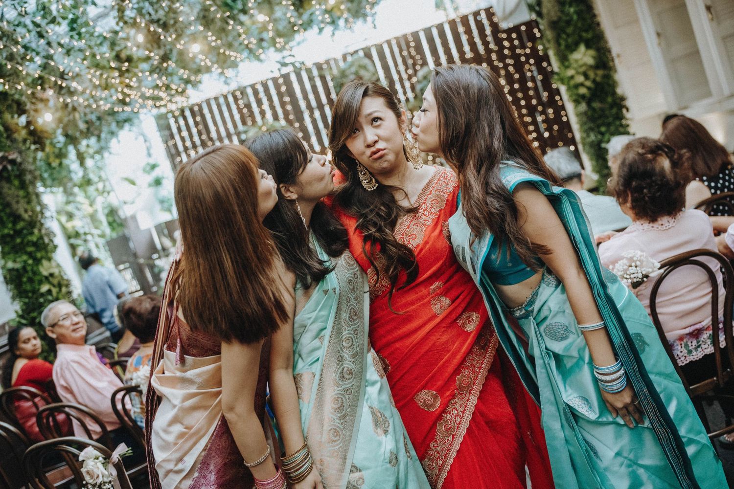 Bridesmaids in teal sarees kissing Indian bride in red saree at outdoor wedding celebration.