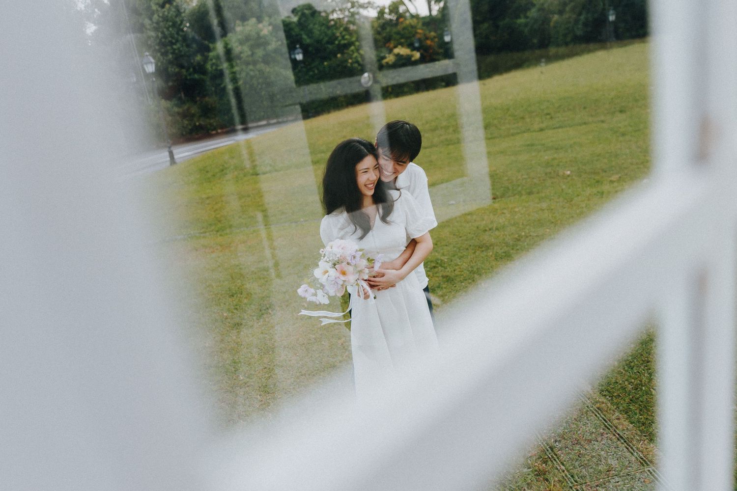 A romantic moment captured through window panes as a couple embraces in white outfits on a grassy lawn.