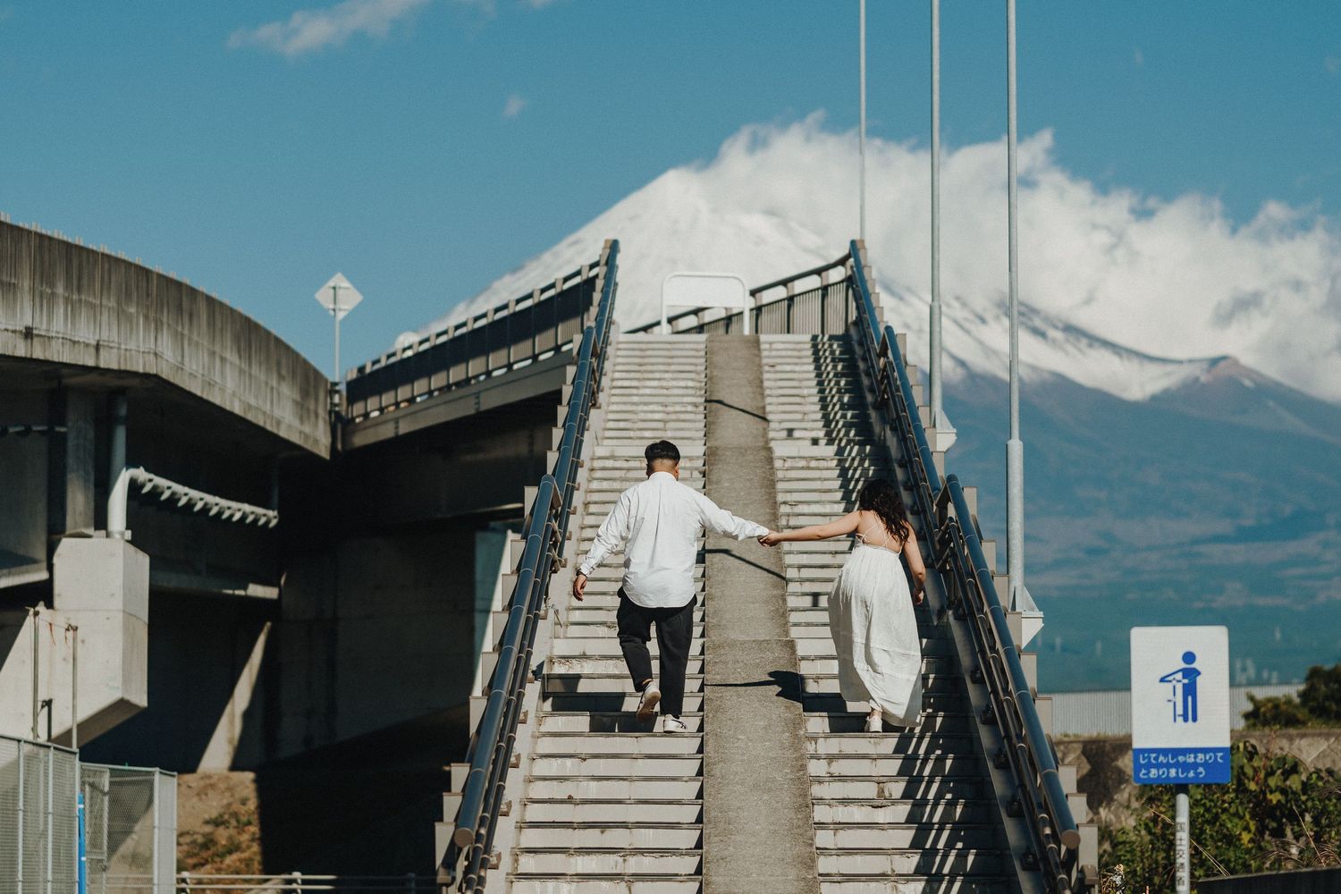 Pedestrian overpass walkway with mountain landscape and blue sky in the background.