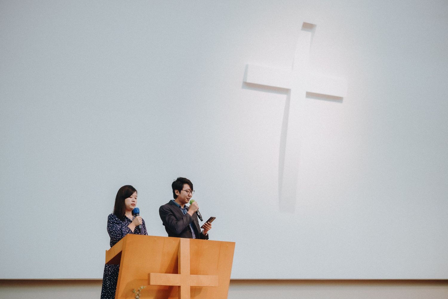 Two people sit in wooden pews beneath a large white cross mounted on a minimalist wall.