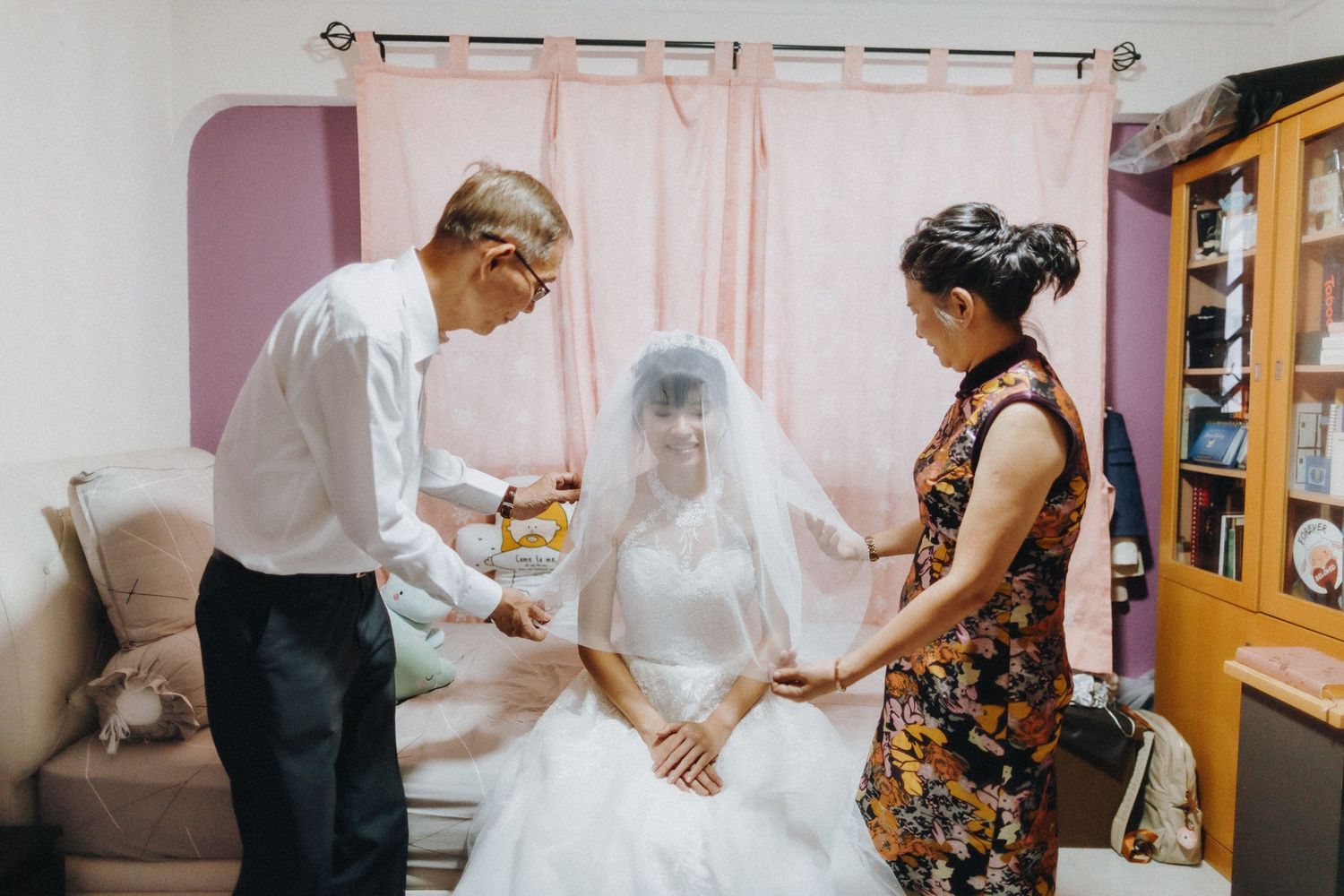 A touching wedding preparation scene shows final adjustments being made to a bride's dress in a pink-tinted room.